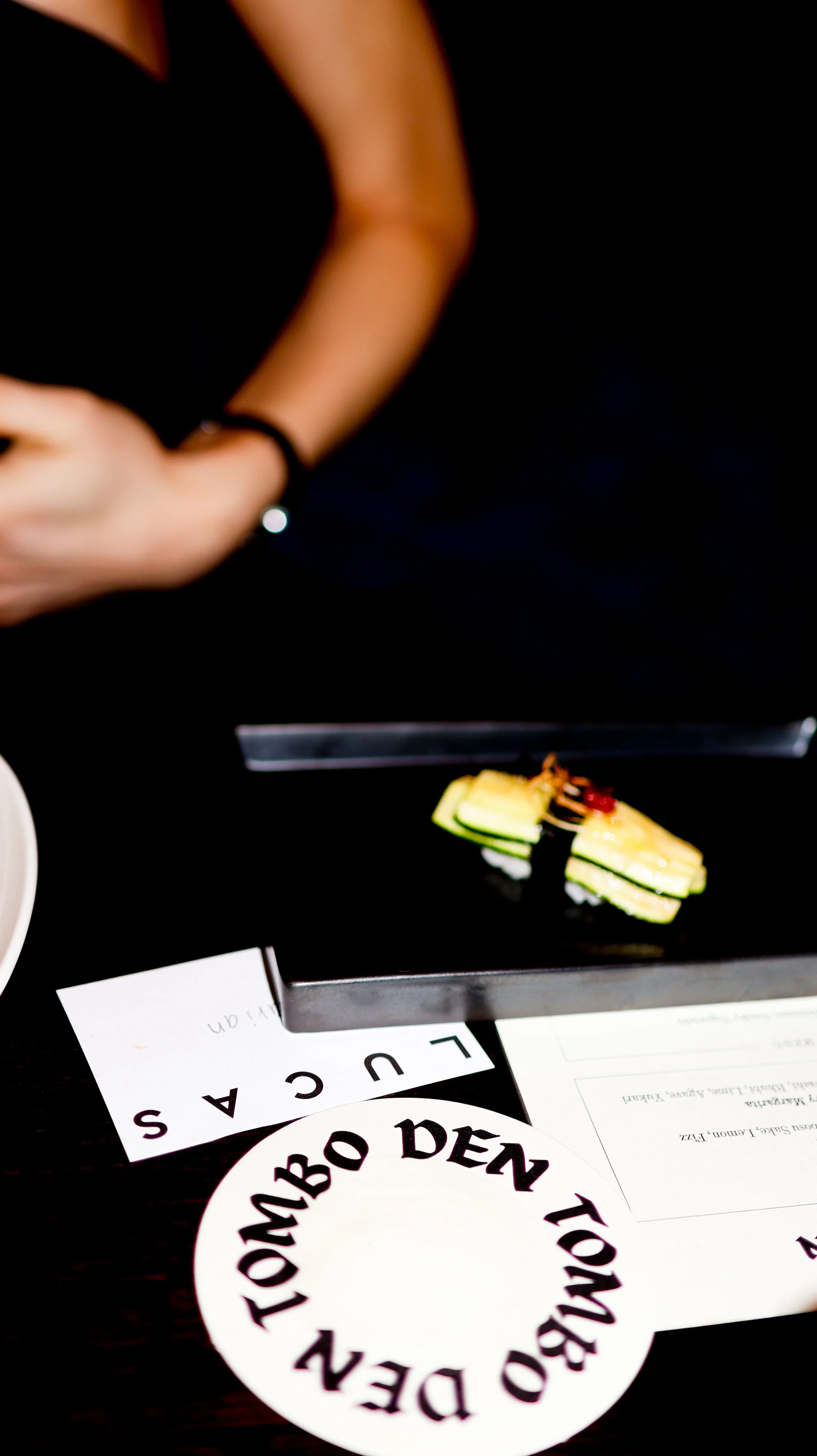 A black tray with a sushi roll and cucumber slices on it, placed on a table with a "Bombay Den TOWN" paper and other paper documents nearby, with a person wearing a black top partially visible in the background.