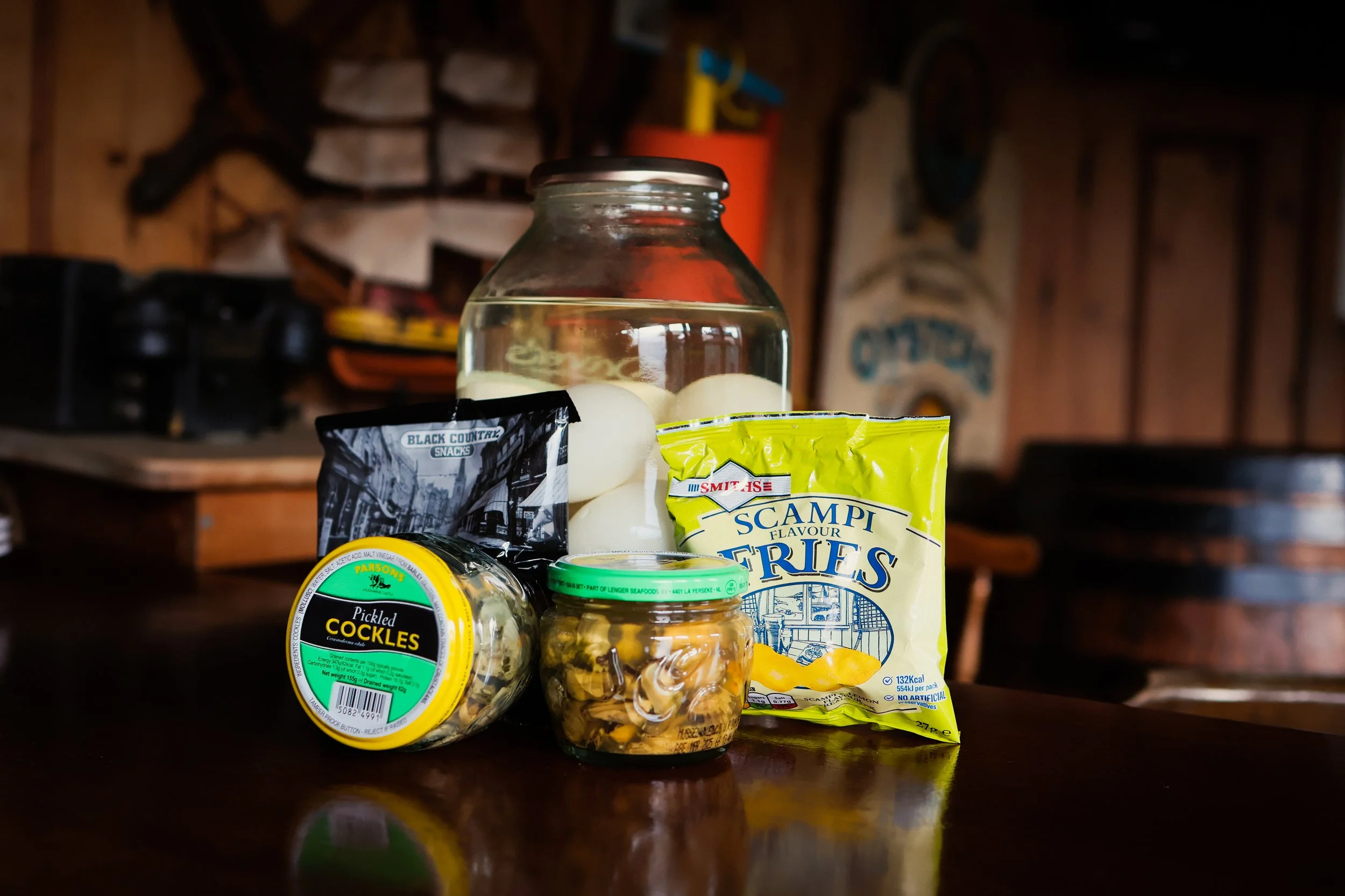 A jar of pickled cockles, a bag of scampi fries, a jar of marinated mushrooms, a bag of scampi fries chips, and a container of pickled boiled eggs on the bar of a pub whose website and social media I manage.