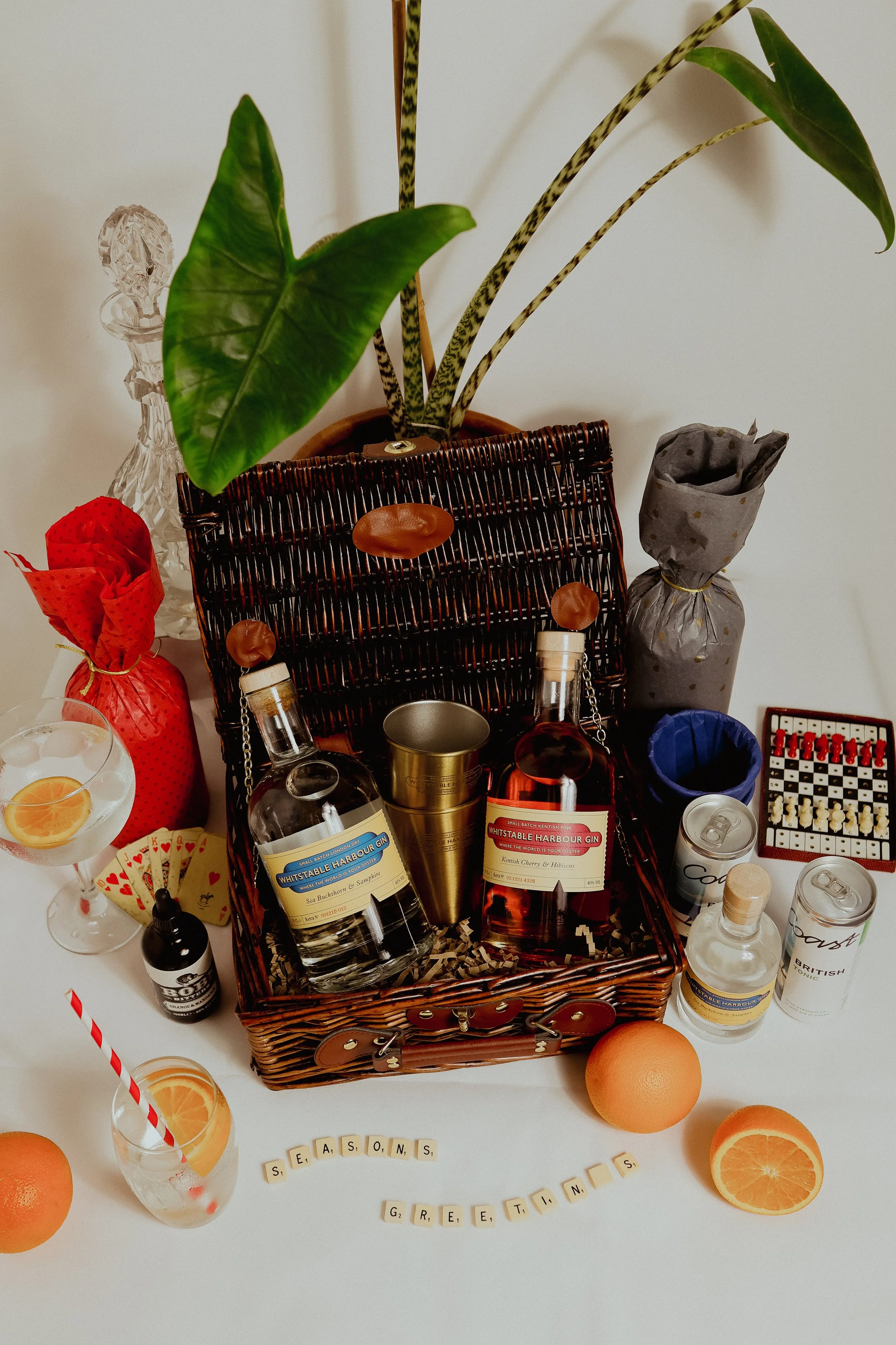A festive arrangement of drinks, gifts, and decorations on a table, with a basket containing bottles of whiskey and gin, oranges, a cocktail with lemon, and various wrapped presents, as part of a Christmas hamper campaign for a client.