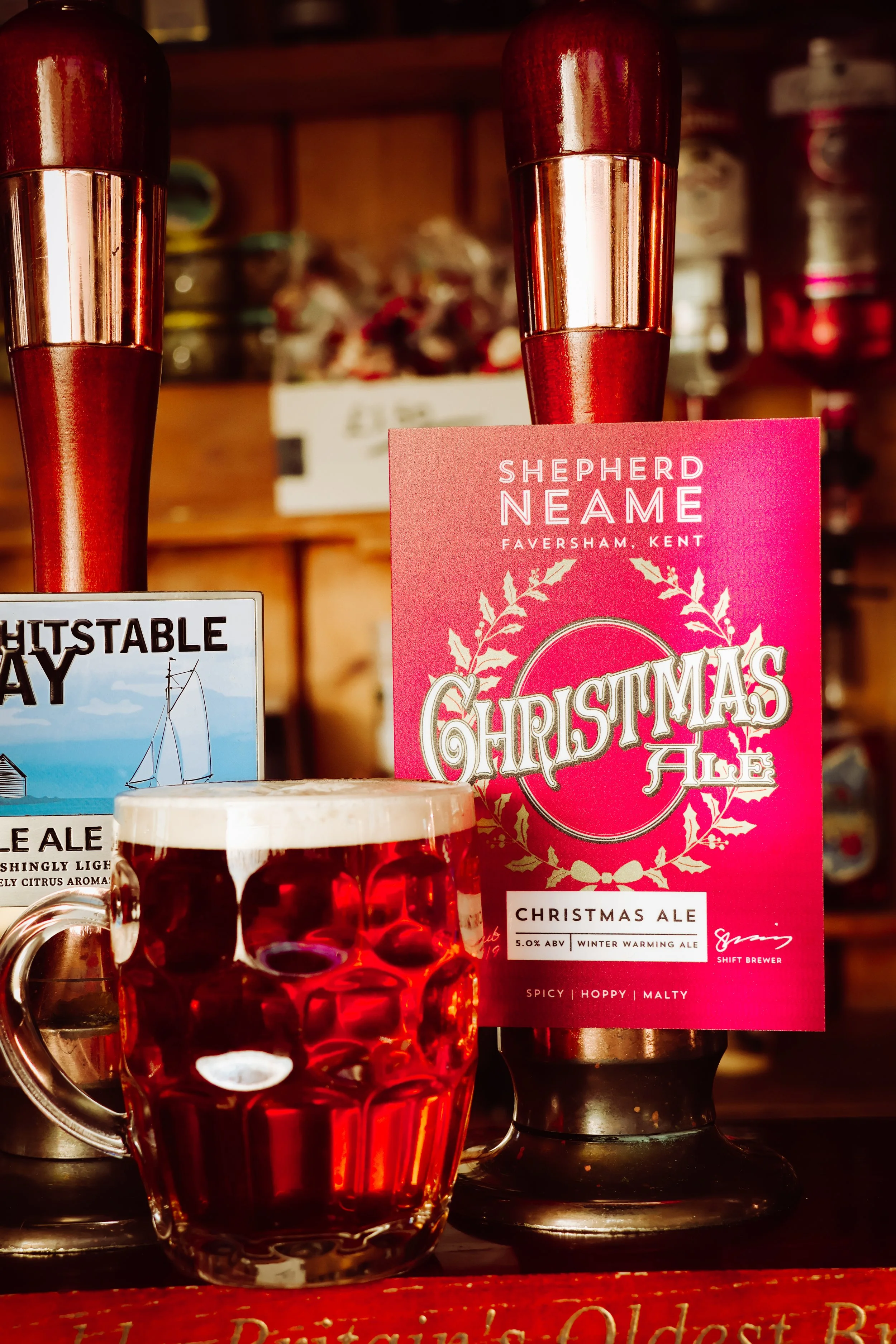 A glass of red Christmas ale with foam on top, a pink promotional card for Shepherd Neame Christmas Ale, and a blue and white sign for Hootstable Bay Ale in the background.