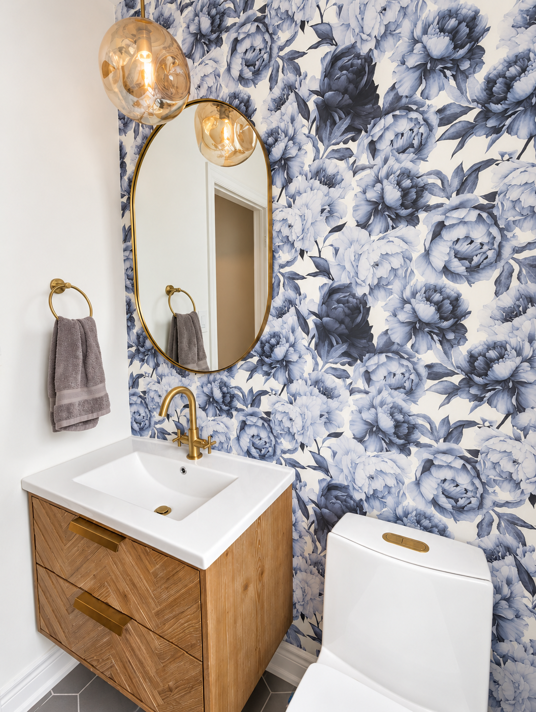 A bathroom with floral patterned wallpaper, a gold-framed mirror, a white sink with a wooden cabinet, a gold faucet, a towel ring with a gray towel, a modern toilet, and a pendant light fixture with a glass shade.