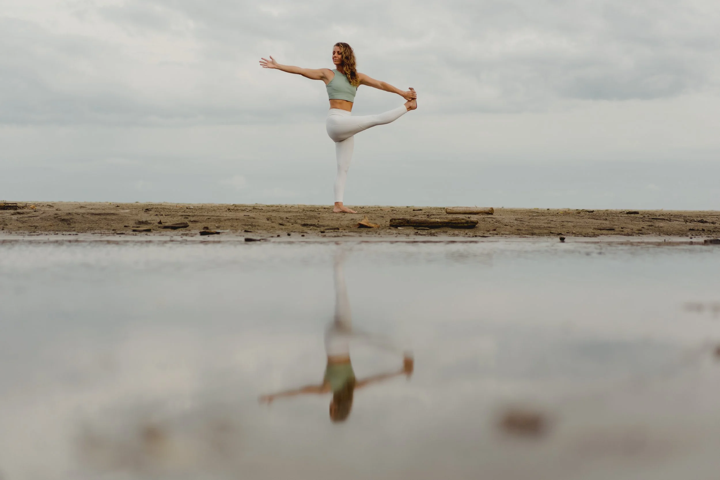 A woman practicing yoga on a sandy beach, balancing on one leg with her arms outstretched, reflection visible in a shallow pool of water.