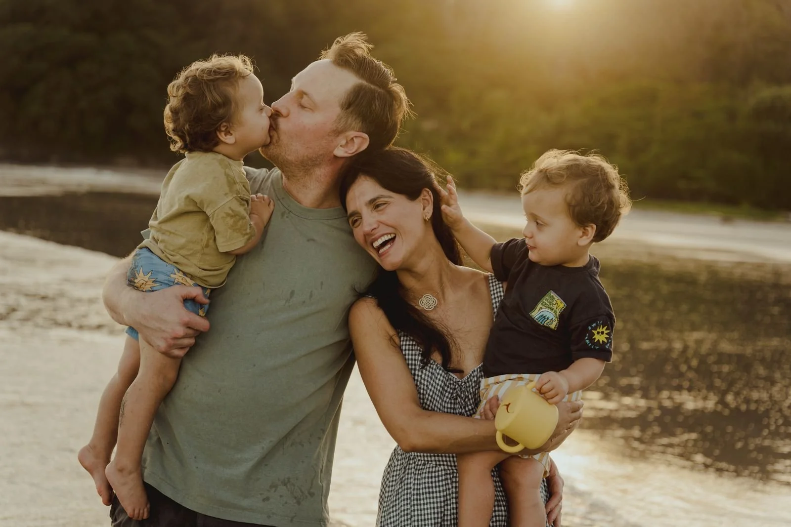 Family outdoors by the water at sunset, father kissing a child, mother smiling holding another child with a yellow cup
