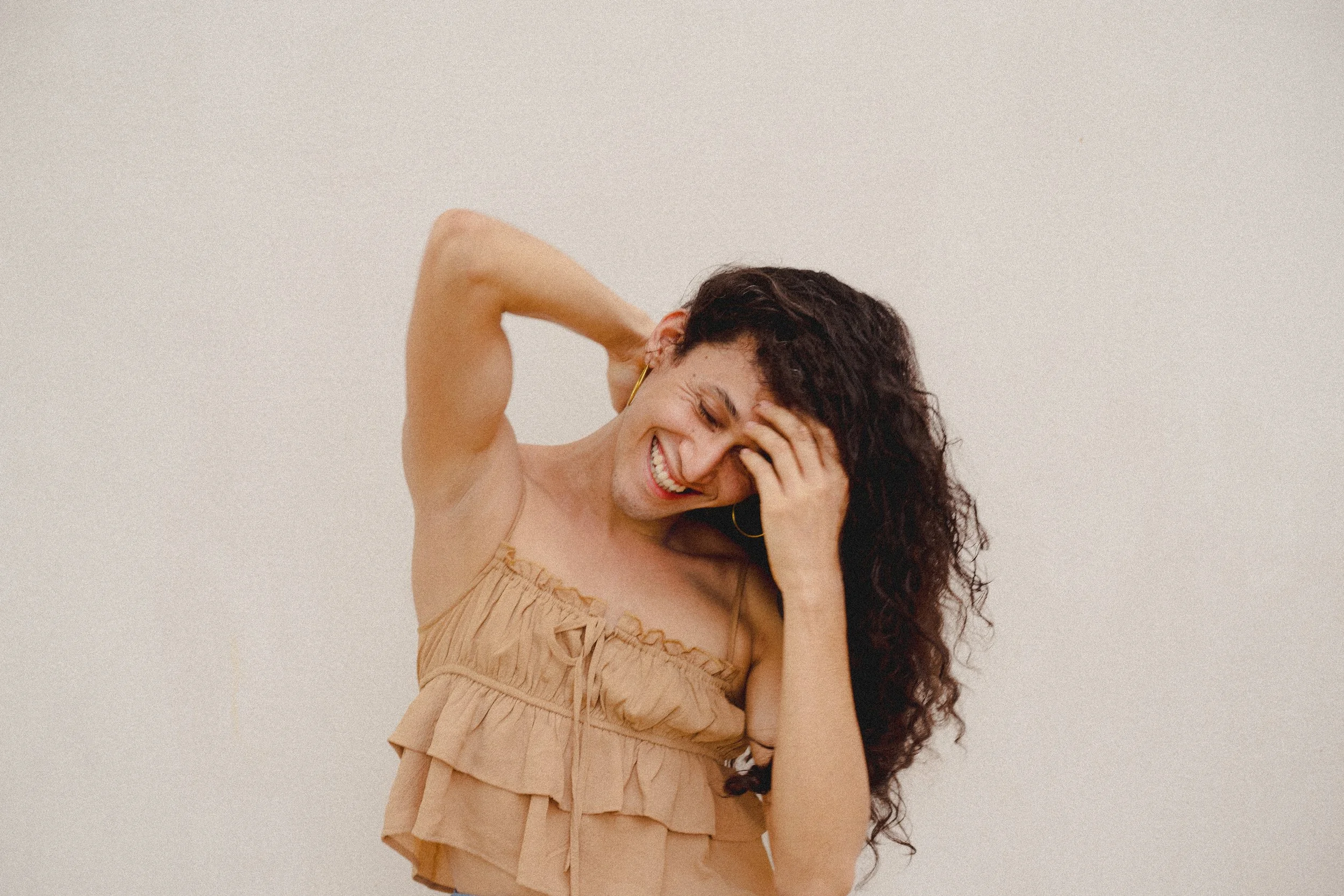 A woman with dark curly hair, wearing a beige ruffled top, laughing and touching her forehead with her hand against a plain light background.