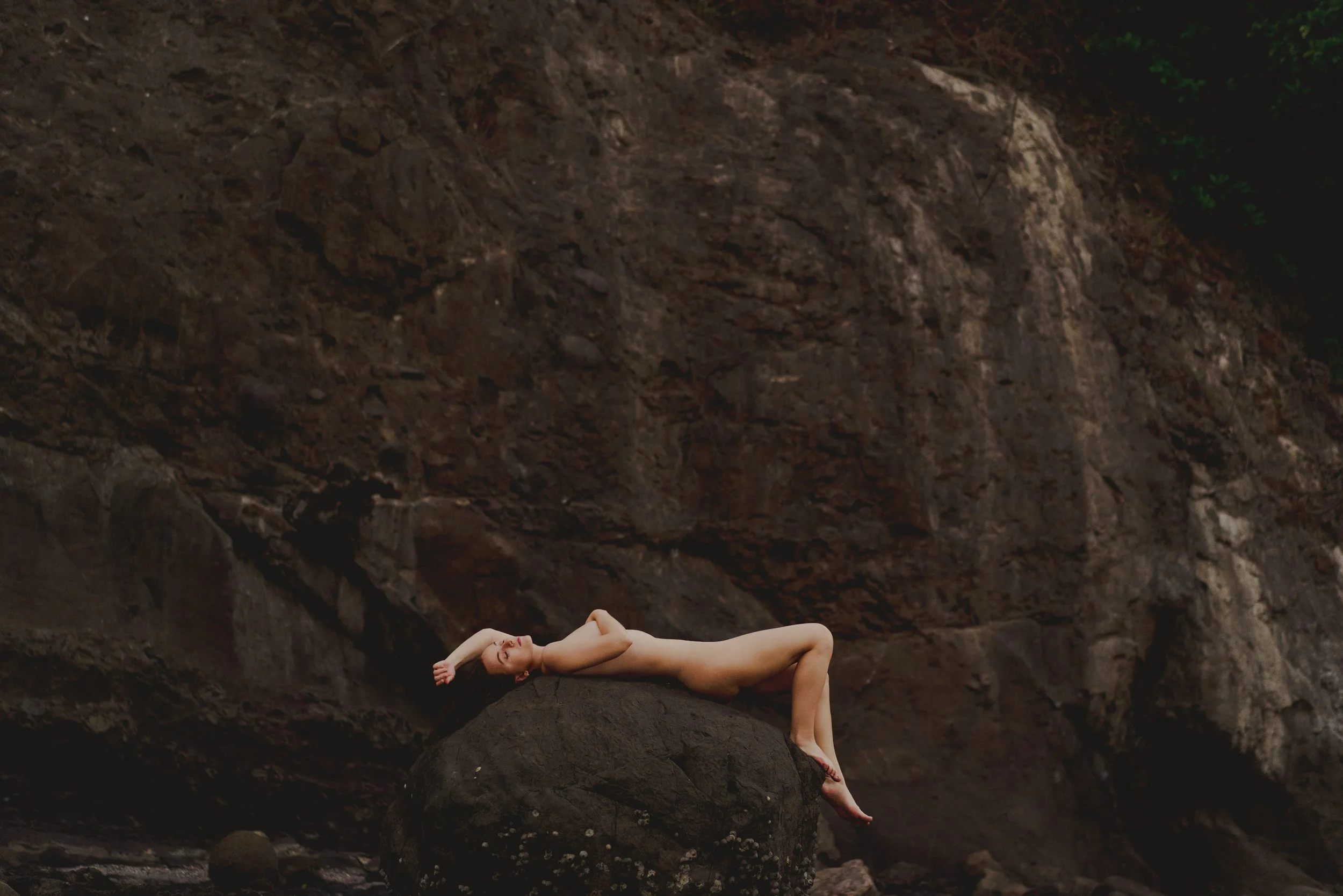 A nude woman lying on a large rock in a natural outdoor setting with large rocky cliffs behind her.