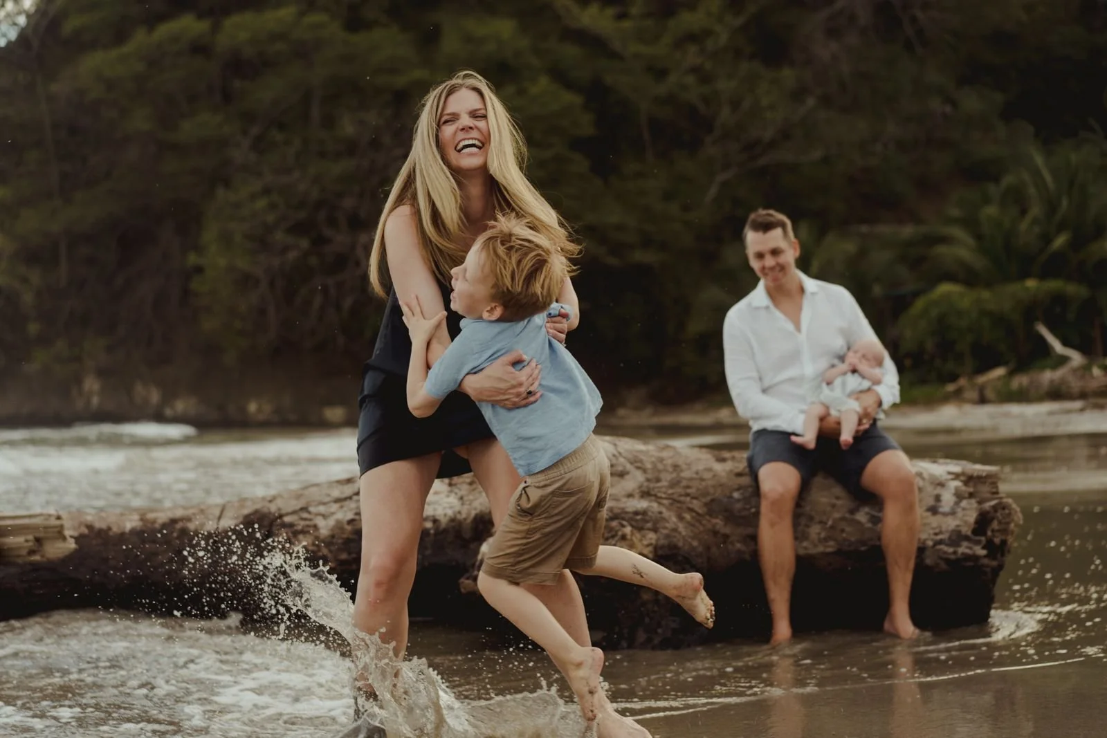 Family enjoying time on a beach, a woman is splashing in the water while laughing, a boy is holding on to her, and a man in the background is sitting on a log holding a baby.