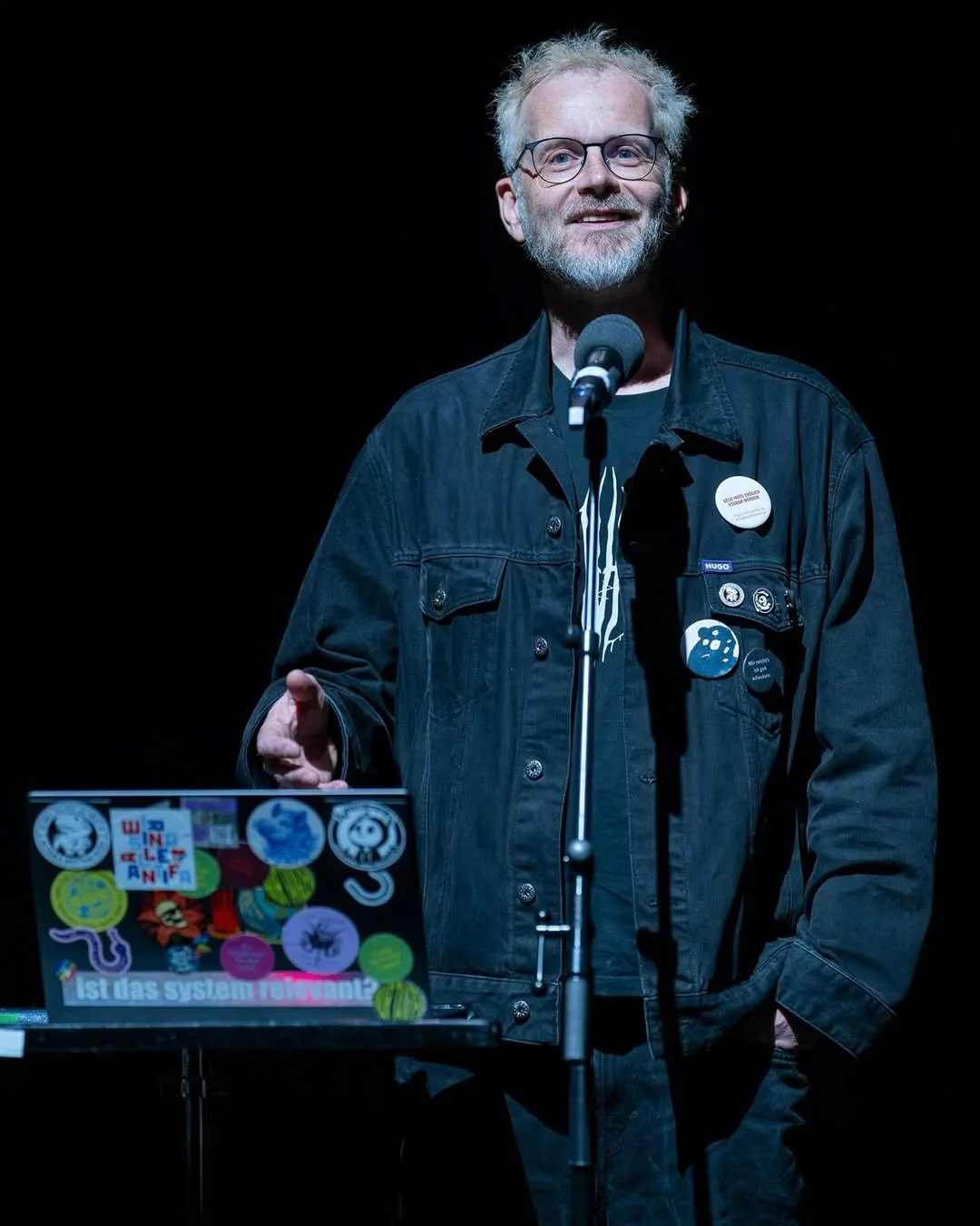 Alex Hellnagel spricht auf der Bühne. Er trägt eine dunkle Jacke und hat mehrere Buttons auf der Jacke. Vor ihm steht ein Laptop, der mit bunten Aufklebern dekoriert ist, und auf dem Tisch liegt ein Mikrofon. Der Hintergrund ist dunkel.