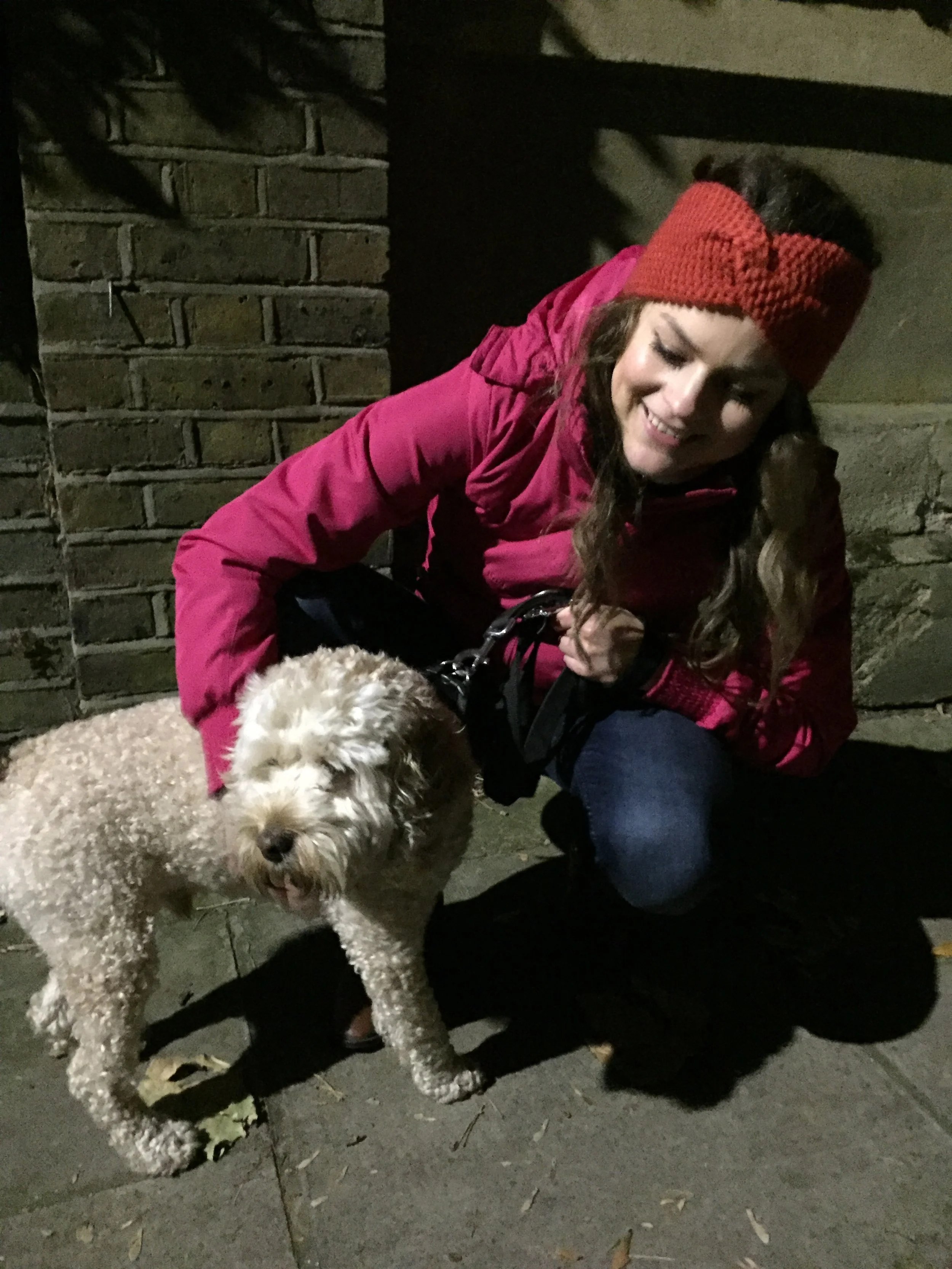 Woman in a pink jacket and orange headband petting a curly-haired dog at night.