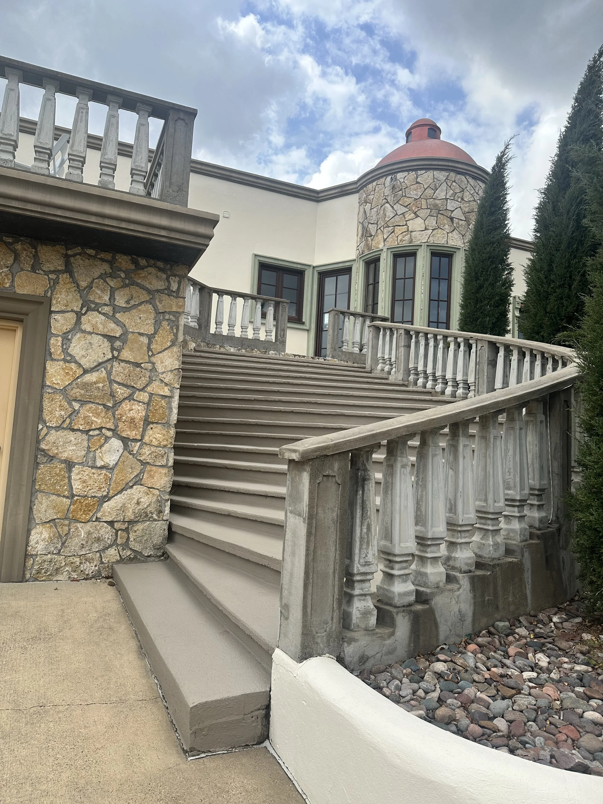 A tuscan style grand outdoors staircase curves up to a Tuscan style villa wedding venue. There is lots of stone. There are tall green trees, and a cloudy sky.