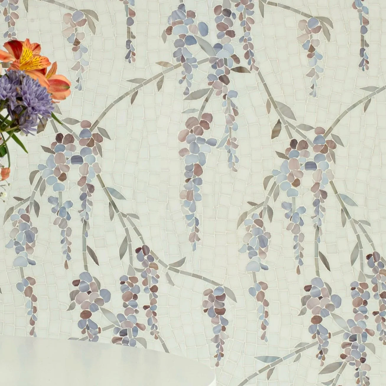 Close-up of a wall with a mosaic of wisteria plant with hanging purple flowers and green leaves, with a small bouquet of mixed colorful flowers on the left side.