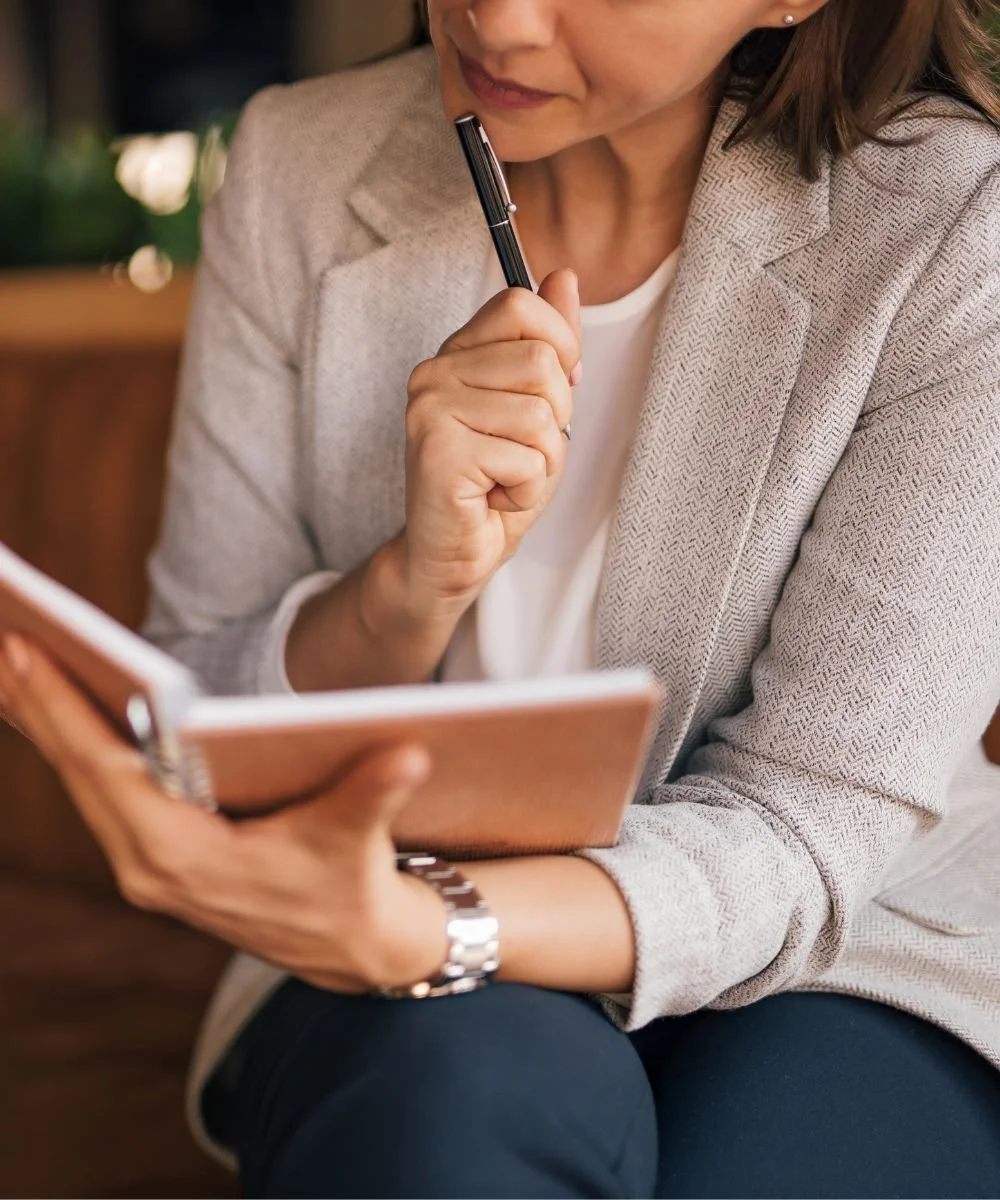 A woman holding a notebook and a pen, appearing to be engaged in reading or writing thoughtfully.