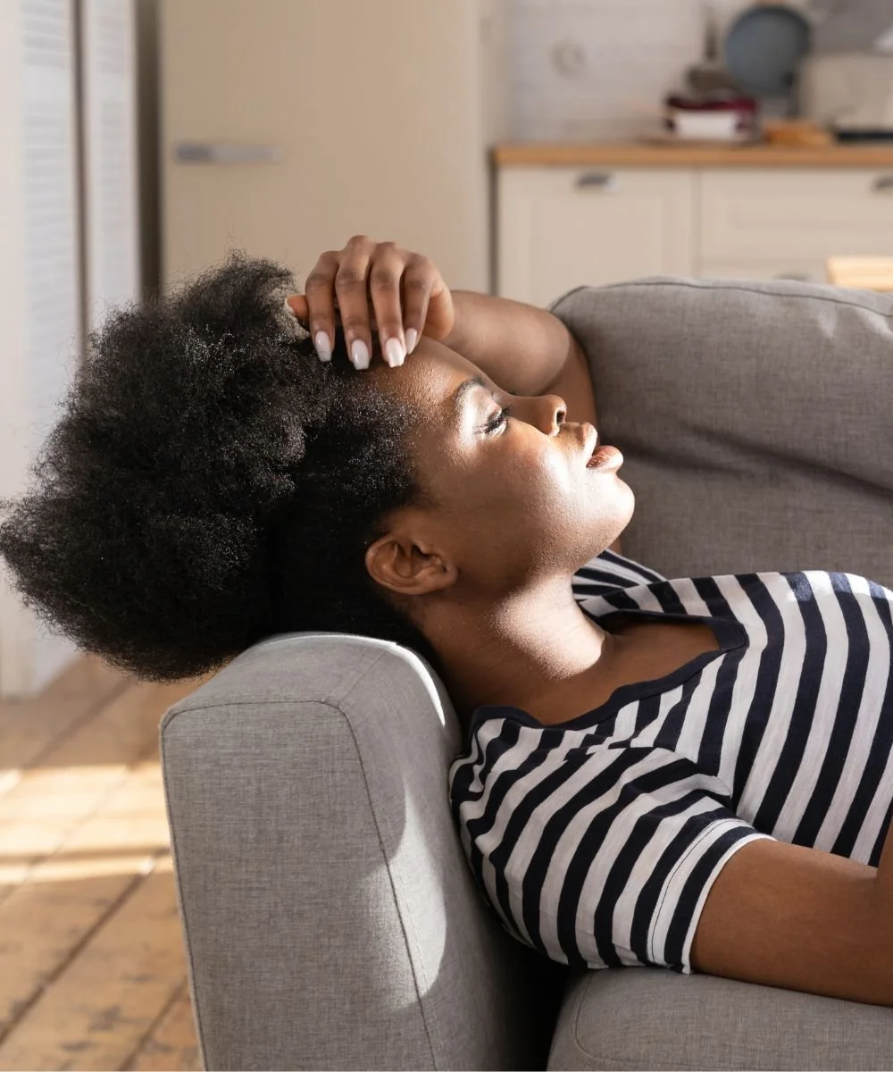Woman with natural curly hair relaxing on a grey couch in a striped shirt, with sunlight shining on her face and hand on her head.
