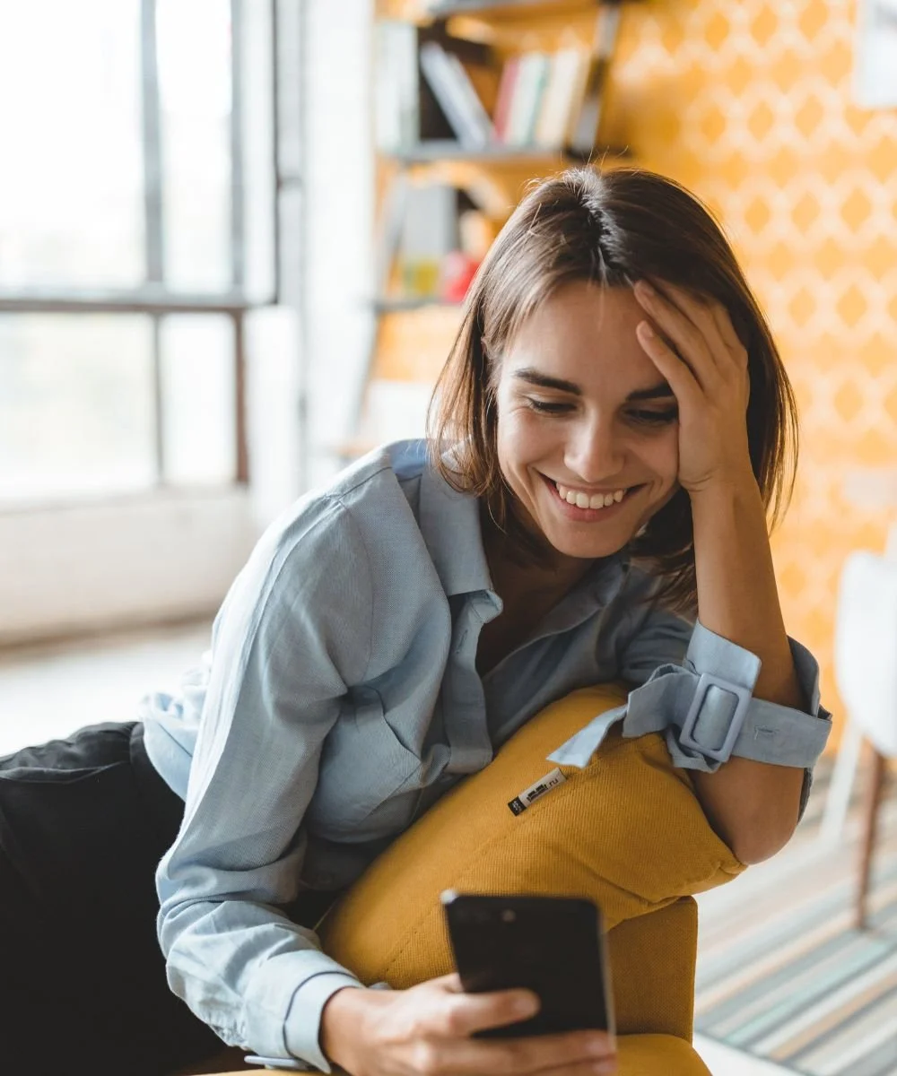 Woman smiling at her phone while sitting on a yellow chair
