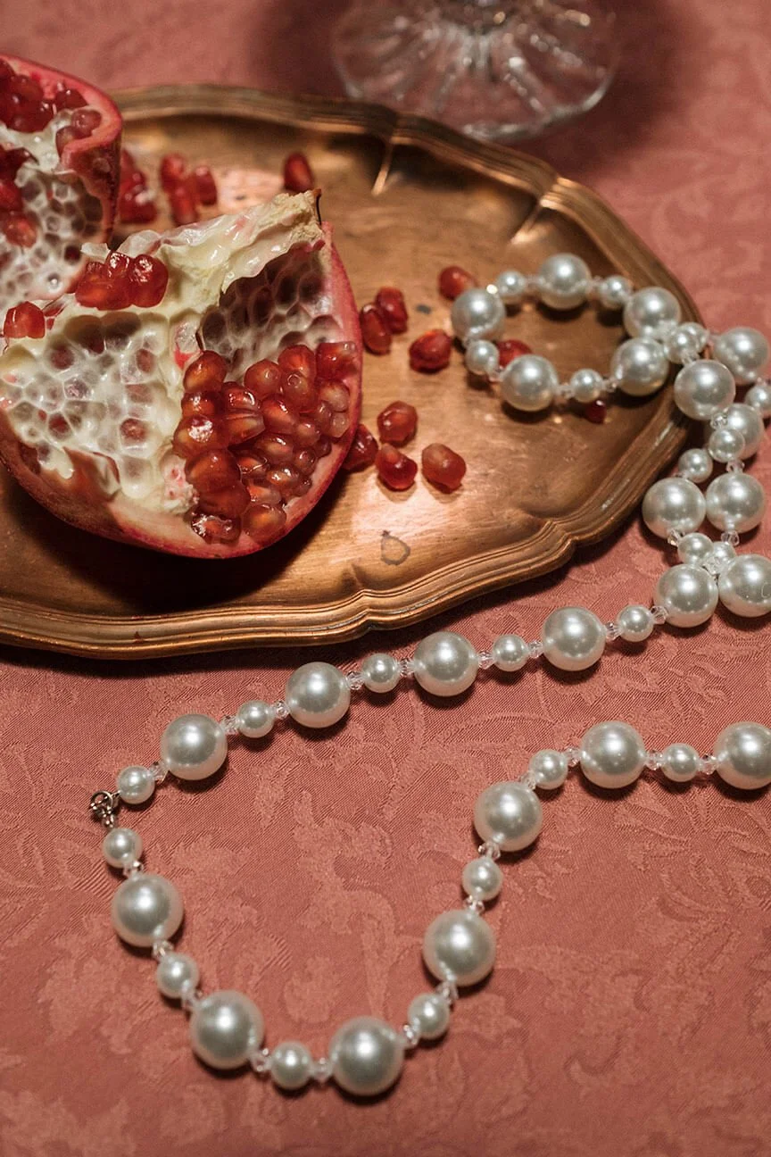 A pomegranate cut open with red seeds on a copper tray, a pearl necklace, and a glass of water on a pink textured tablecloth.