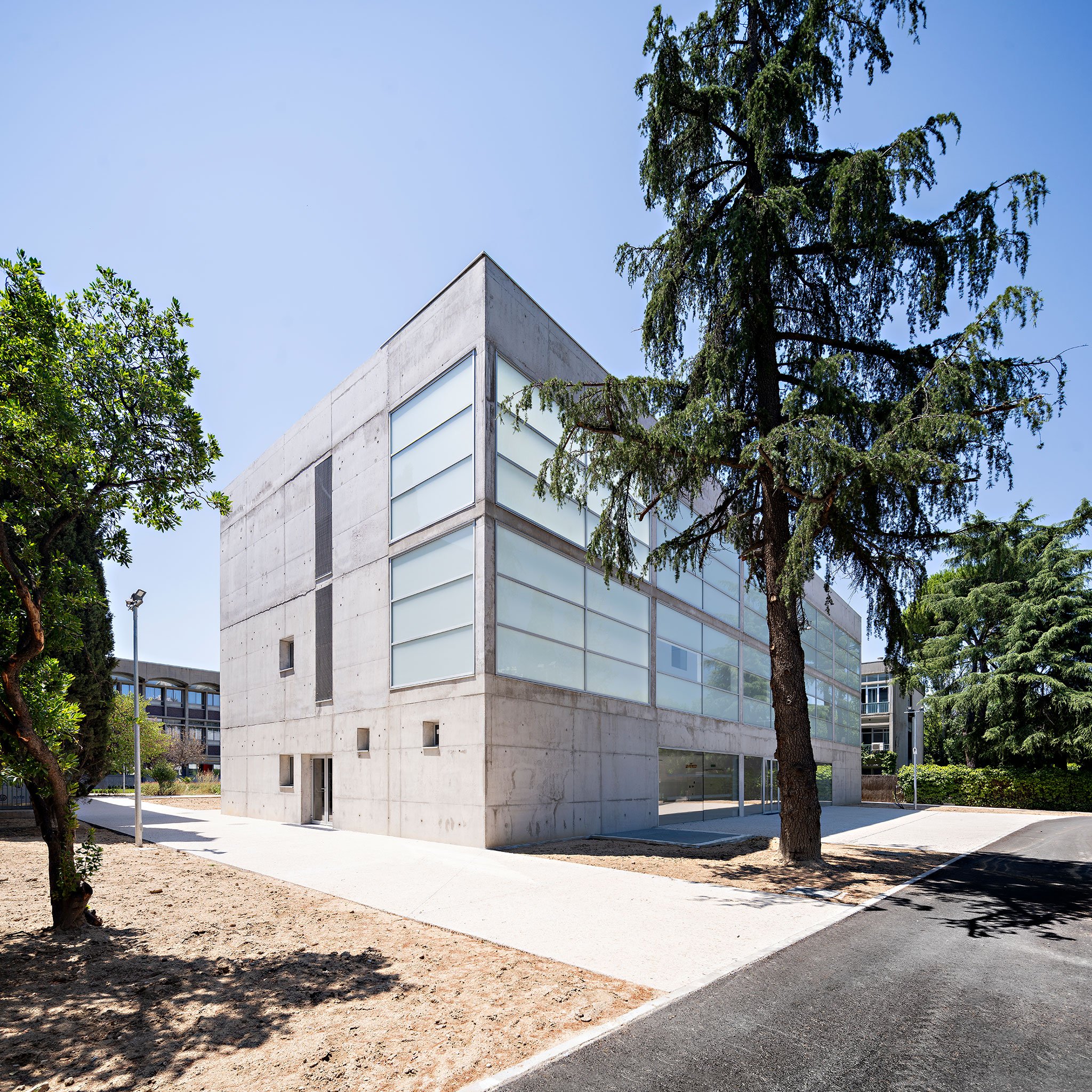 Modern concrete building with large glass windows, surrounded by trees and a paved pathway.