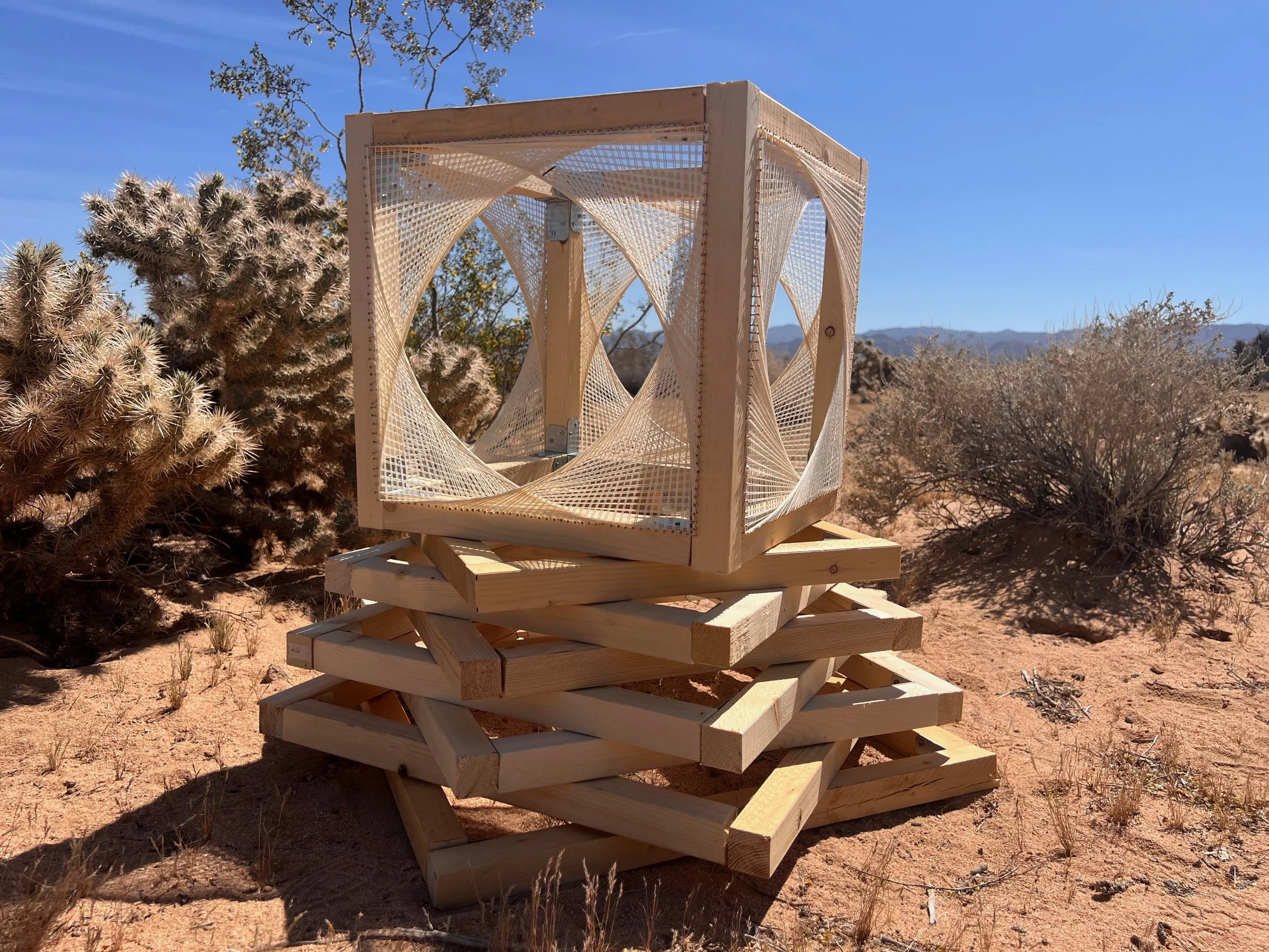 A wooden geometric outdoor sculpture with fabric inserts, placed on a layered wooden platform in a desert landscape with cacti and shrubs under a clear blue sky.