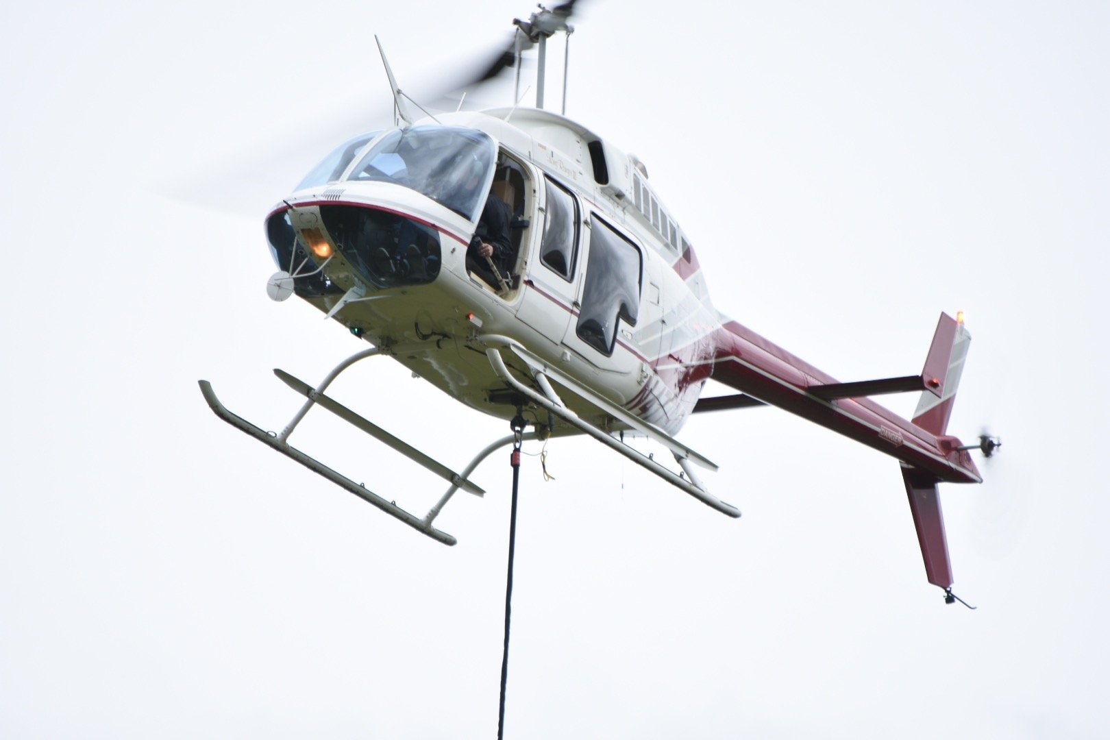Aerial view of a helicopter in flight with a rope hanging below, against a cloudy sky.
