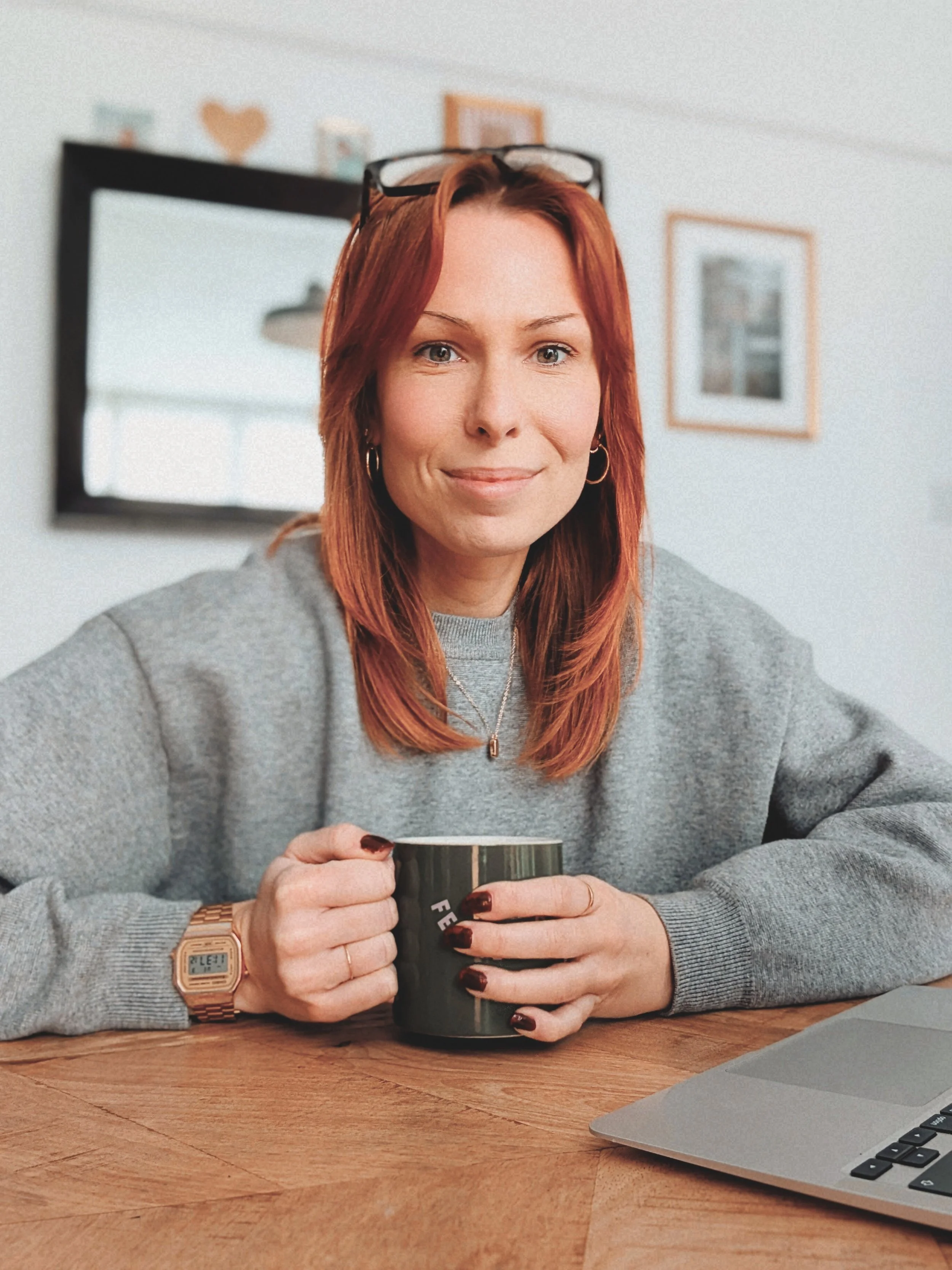 A woman with red hair and glasses on her head sitting at a wooden table, holding a black mug and smiling, with a laptop in front of her and a mirror and framed pictures on the wall behind her.