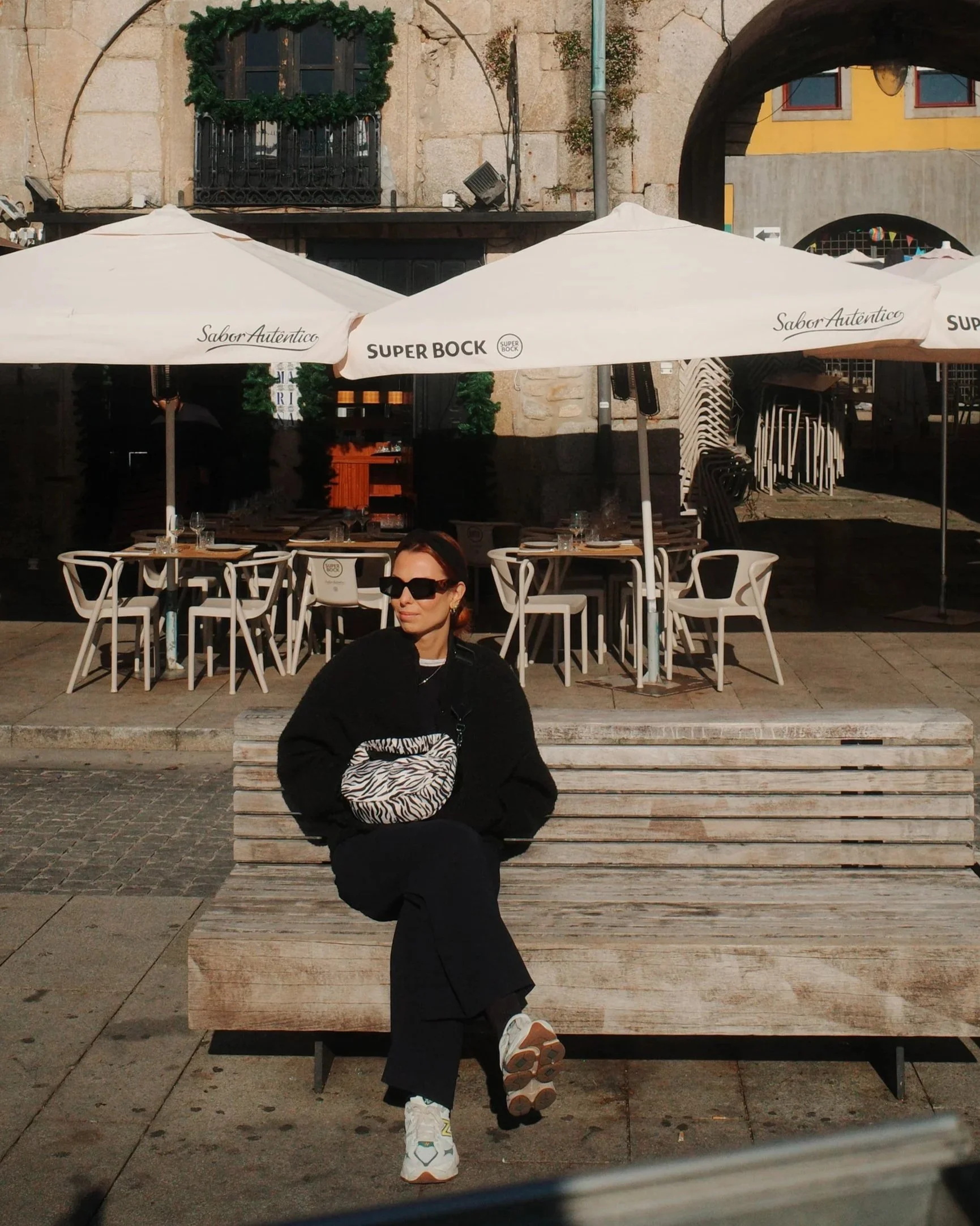 A woman sitting on a wooden bench in an outdoor café area. She is wearing sunglasses, a black coat, and white sneakers, and carrying a zebra-patterned bag. Behind her are white chairs, tables set with glasses, large white umbrellas, and a building with stone walls and an arched opening.