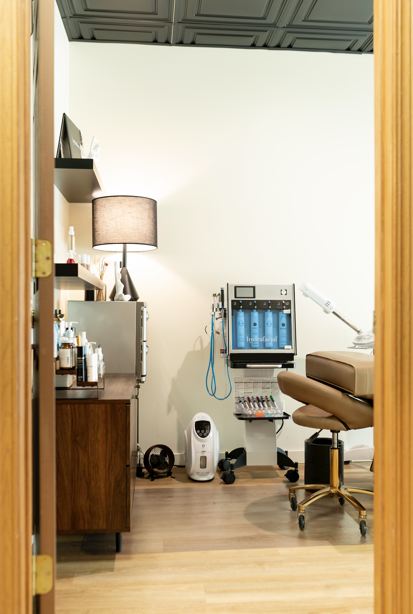 A treatment room with skincare products on a wooden cabinet, a lamp, a small mirror, and a shelf. There are equipment including a skincare machine labeled "hydrafacial," a magnifying lamp, a white air purifier, and a tray of syringes. The room has light-colored walls and a wooden floor with a brown treatment table.