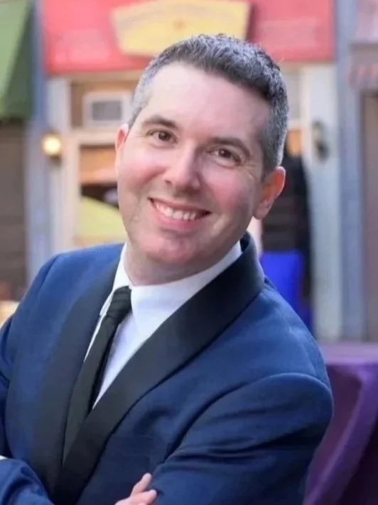 A man in a blue suit with a black lapel, white shirt, and black tie, smiling with arms crossed, standing outdoors in front of a building with signage and umbrellas.