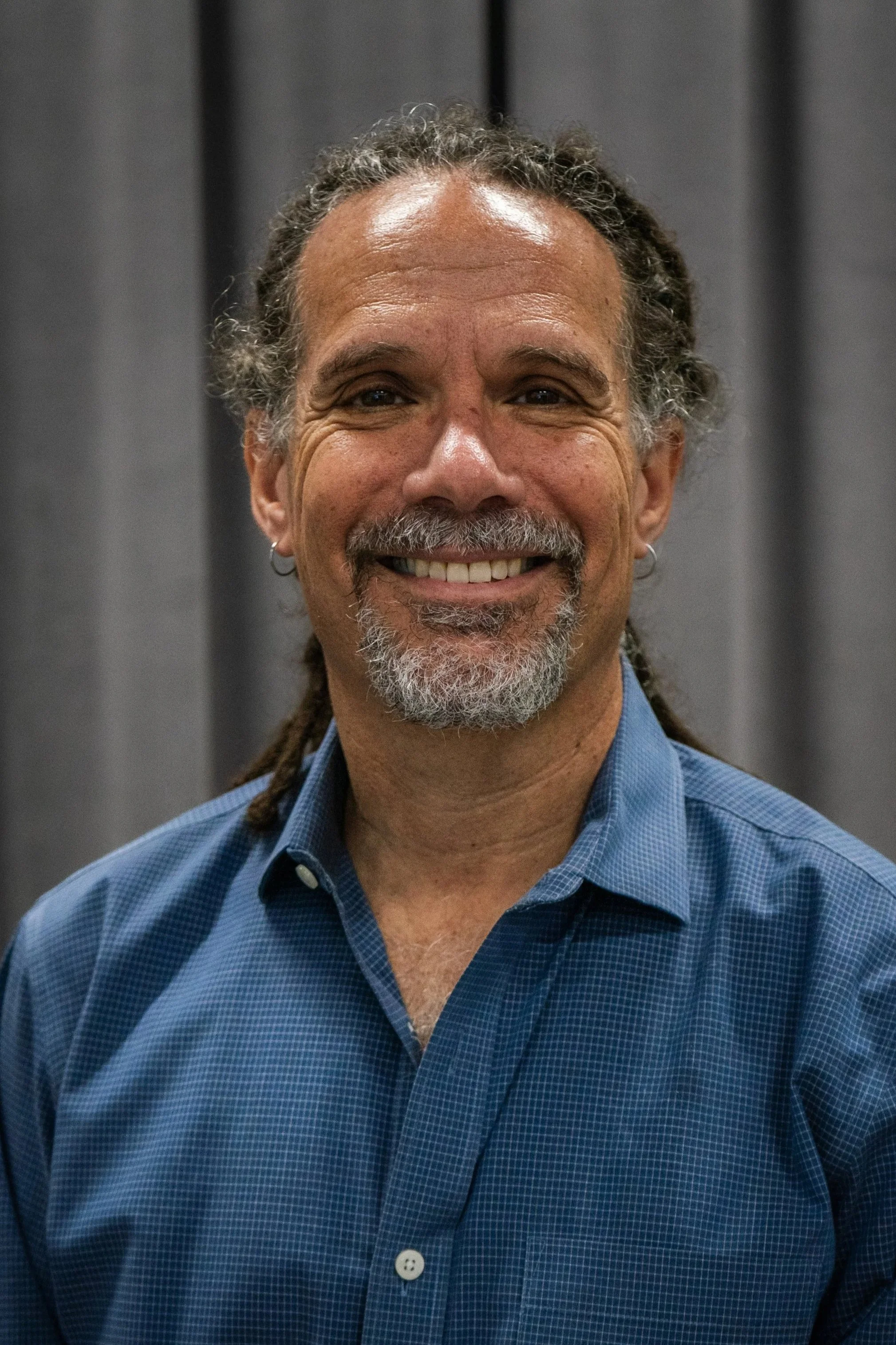 A smiling man with gray curly hair and a beard, wearing a blue checkered shirt, standing in front of a dark gray curtain.