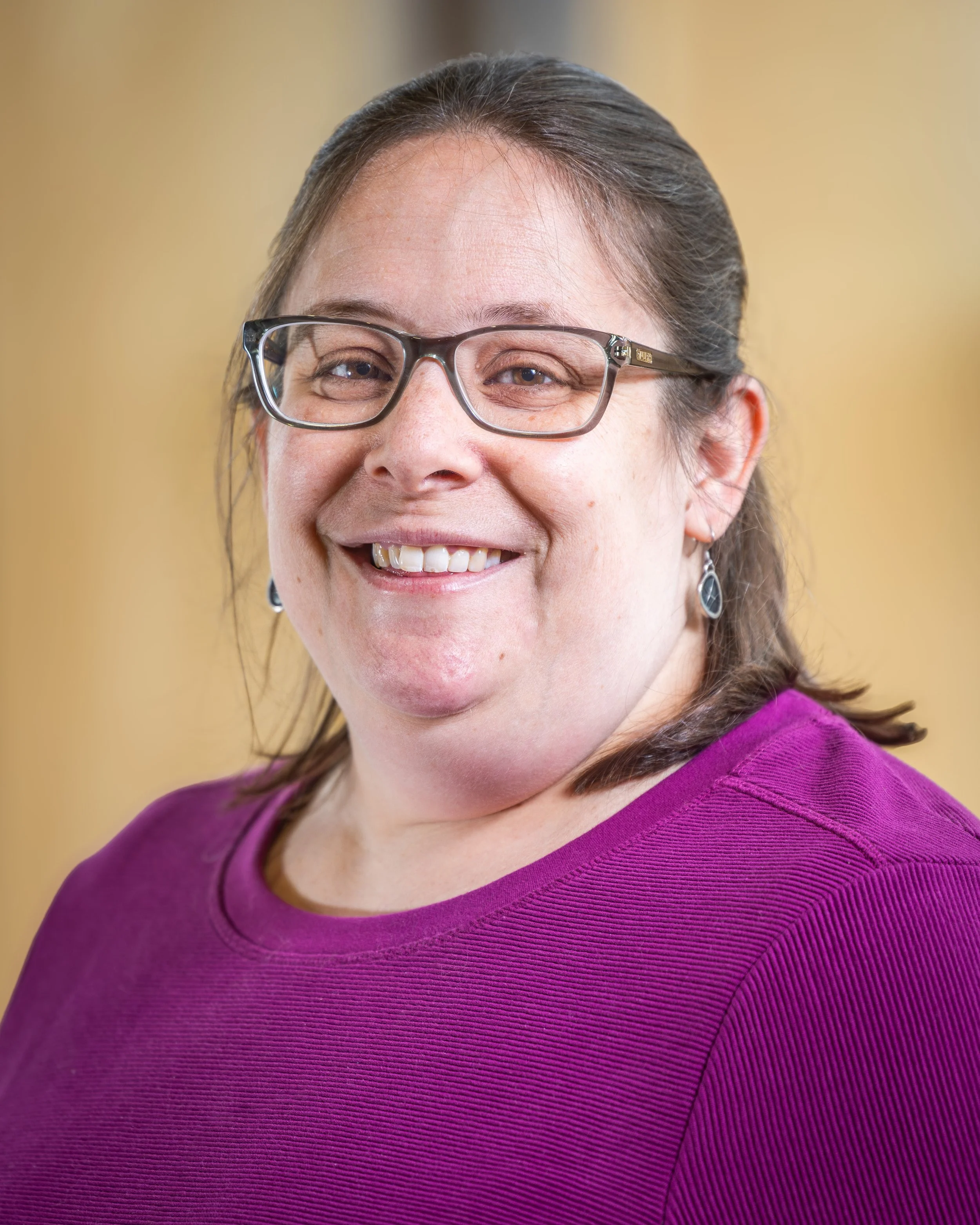 A lady with shoulder length brown hair, glasses, and a purple shirt.