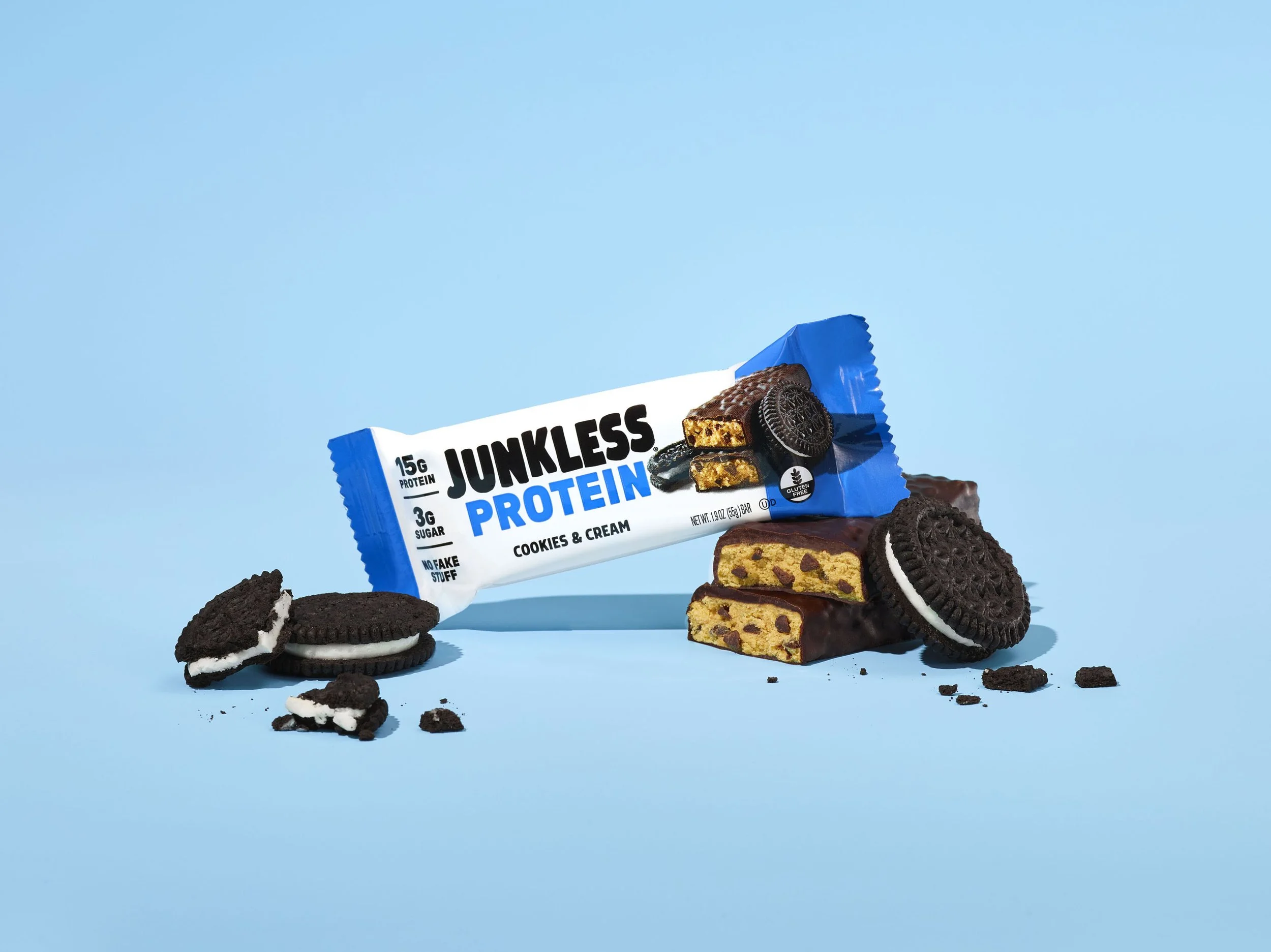 A protein bar labeled "Junkless Protein" with cookie and cream flavor, surrounded by Oreo cookies and broken pieces on a light blue background.