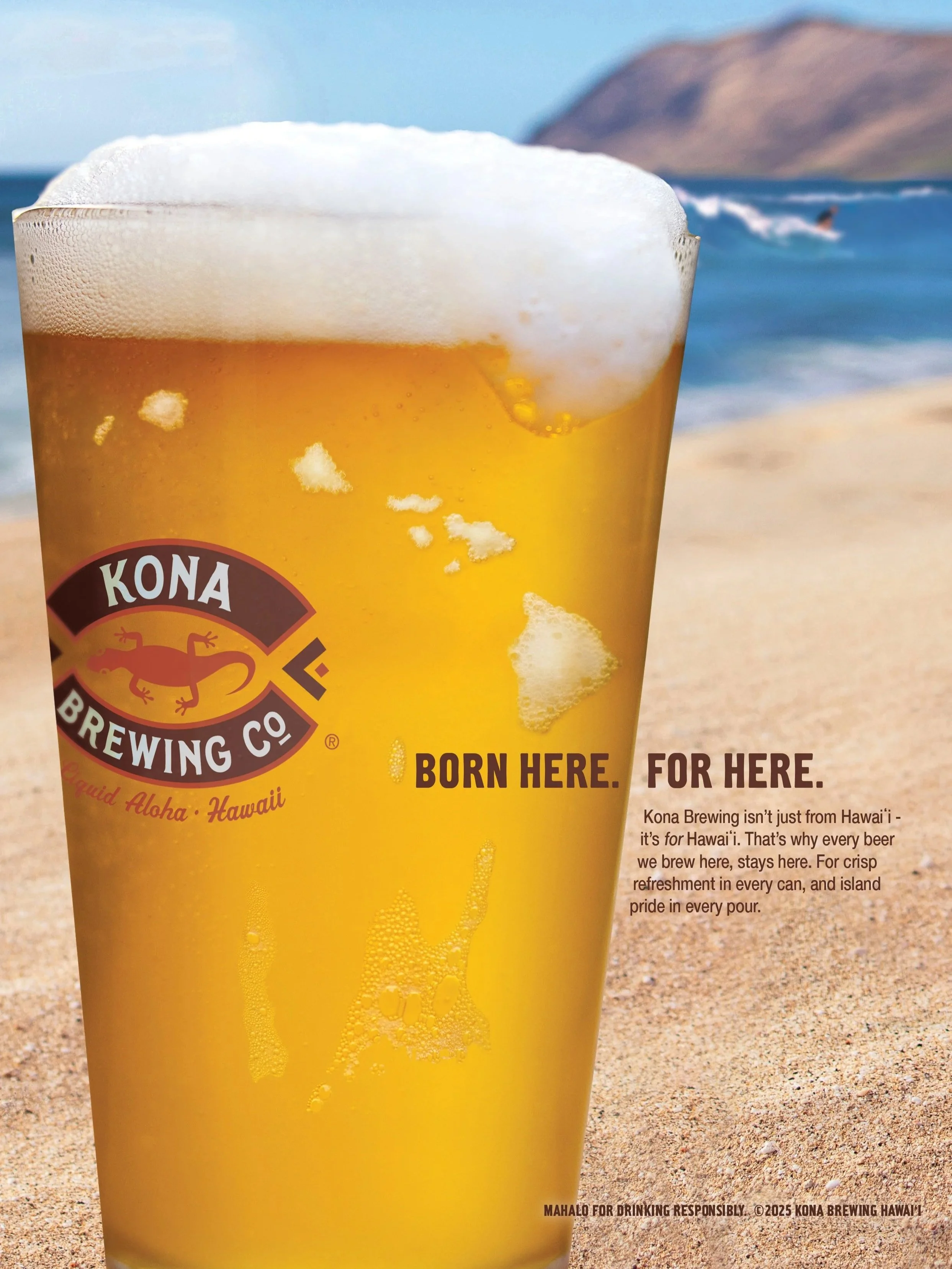 A glass of beer with foam on top, placed on a sandy beach with the ocean and mountains in the background.