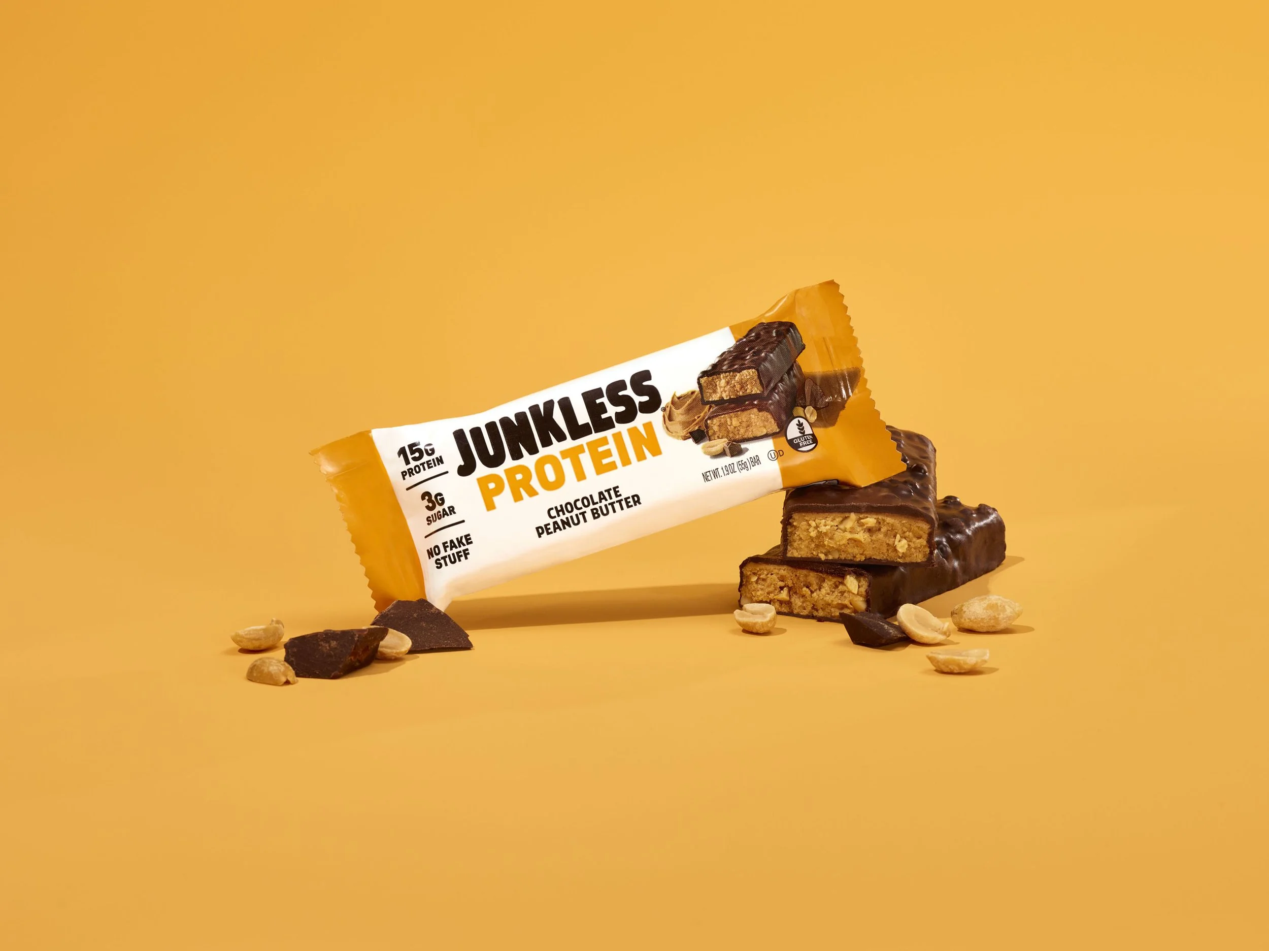 A protein bar labeled 'Junkless Protein' with chocolate and peanut butter flavor, placed on a yellow background with scattered peanuts and chocolate pieces.
