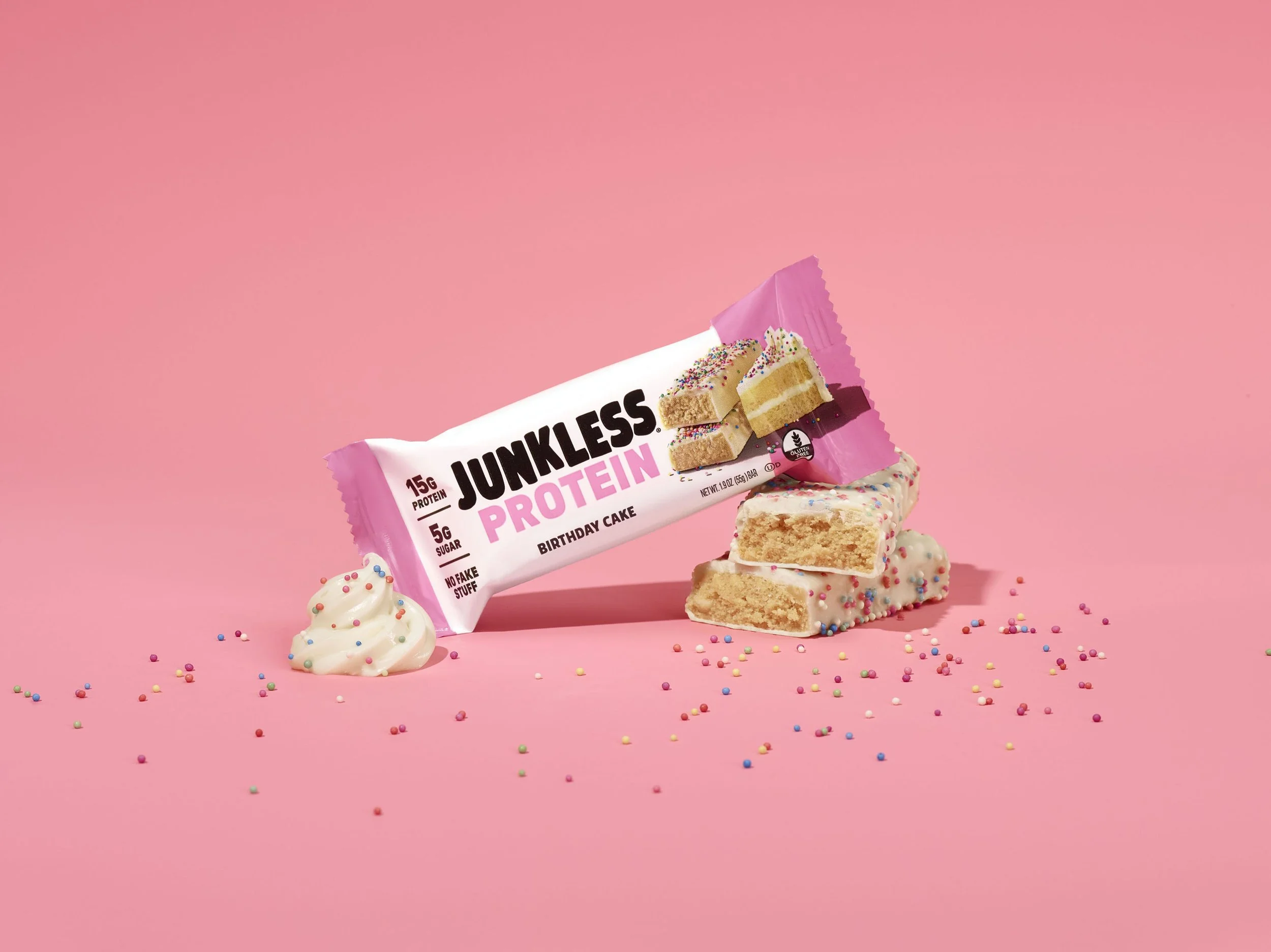 A pink snack bar wrapper labeled 'Junkless Protein' placed on a pink background with pieces of cake and white frosting decorated with multi-colored sprinkles, and scattered sprinkles around.