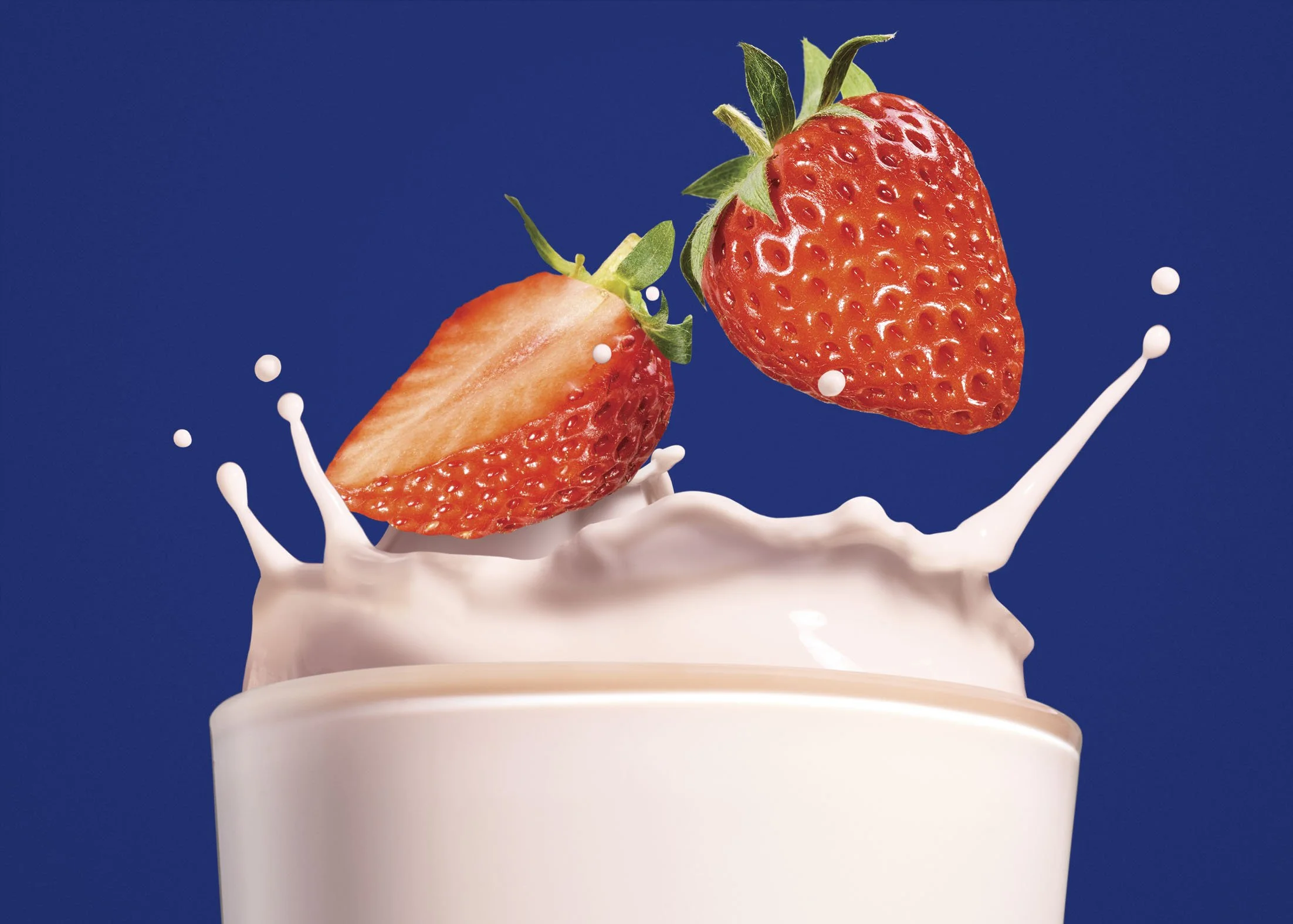 Close-up of a strawberry half and whole strawberry falling into a splash of milk against a dark blue background.