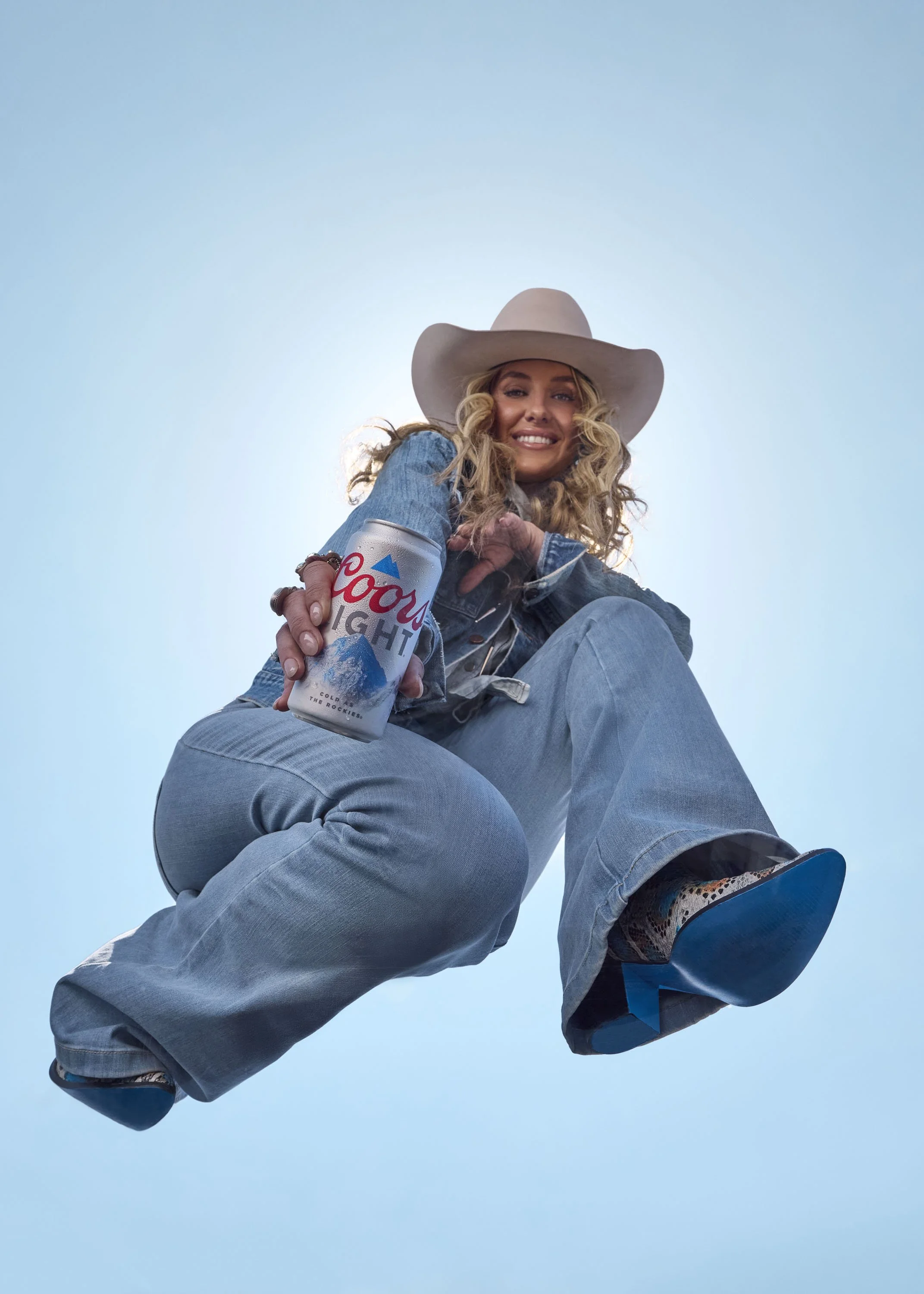 A woman with curly blonde hair wearing a wide-brimmed hat, denim jacket, and jeans, smiling while holding a Coors Light beer can, viewed from below against a clear blue sky.