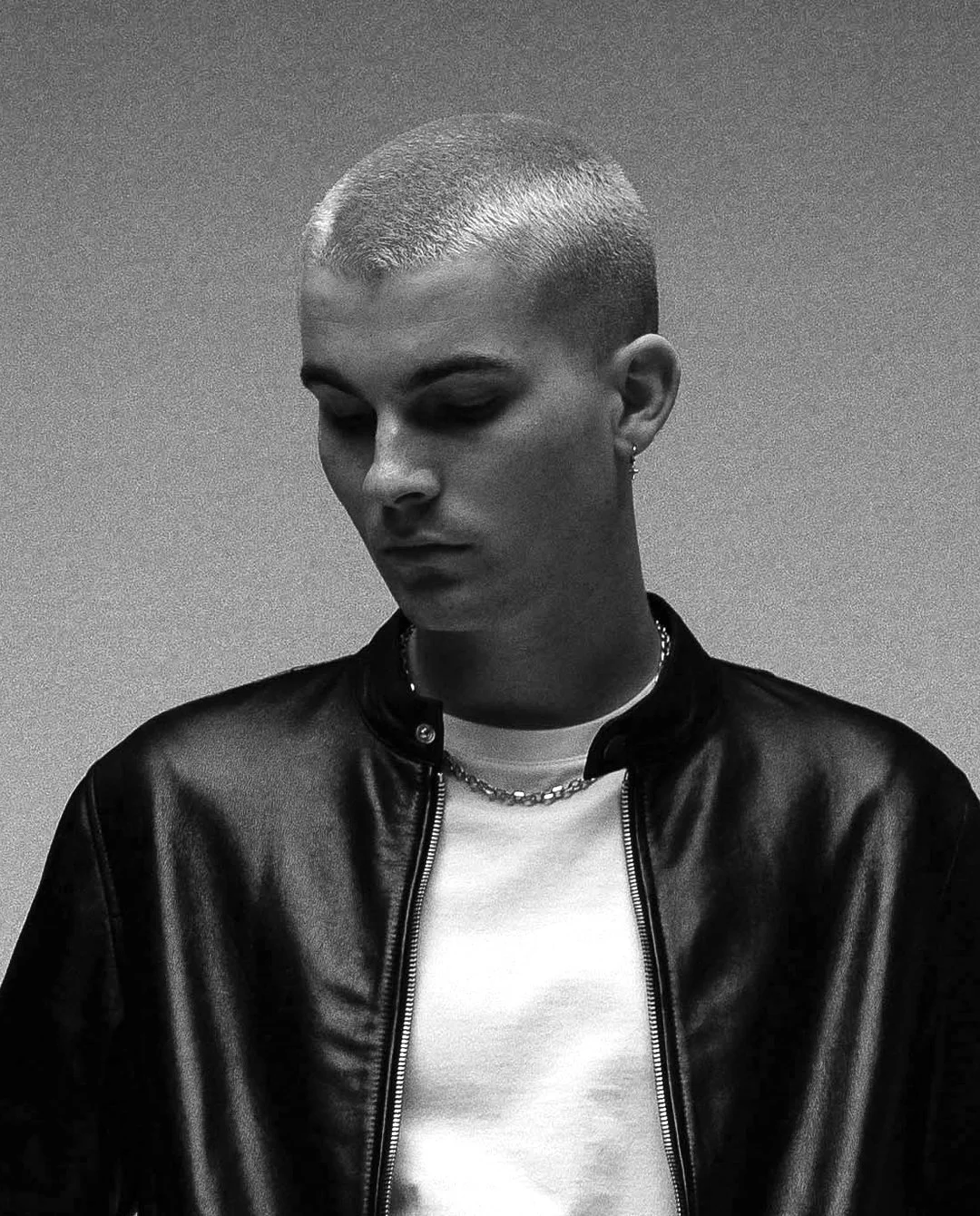 Black and white photo of a young woman with a shaved head wearing a leather jacket, white shirt, and silver jewelry, looking downward.
