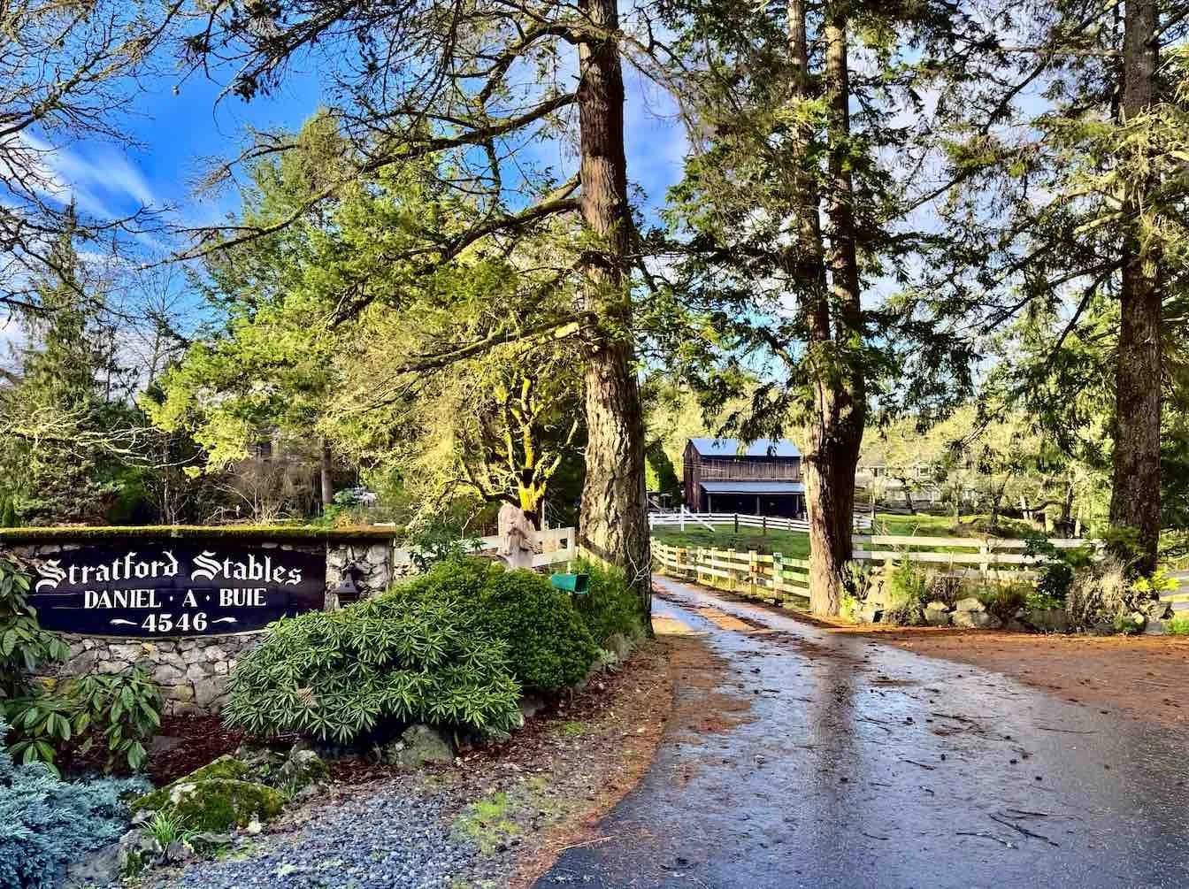 Entrance to Stratford Stables with a driveway, large trees, a sign with the name, and a small barn or stable building in the background.