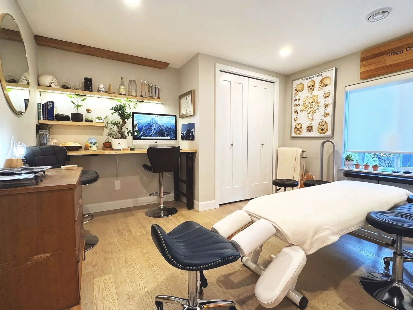 A room with a massage table, a computer desk with a monitor, and various shelves with books and decorative items, under warm lighting and greenery.