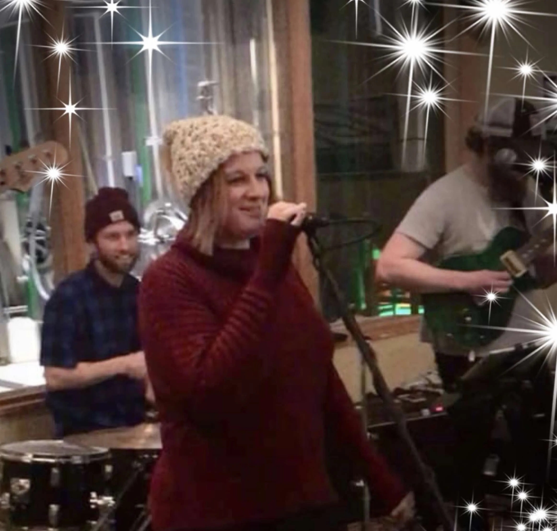 A woman singing into a microphone while a man plays a guitar and another man stands behind them in a cozy indoor setting with sparkling star effects added.