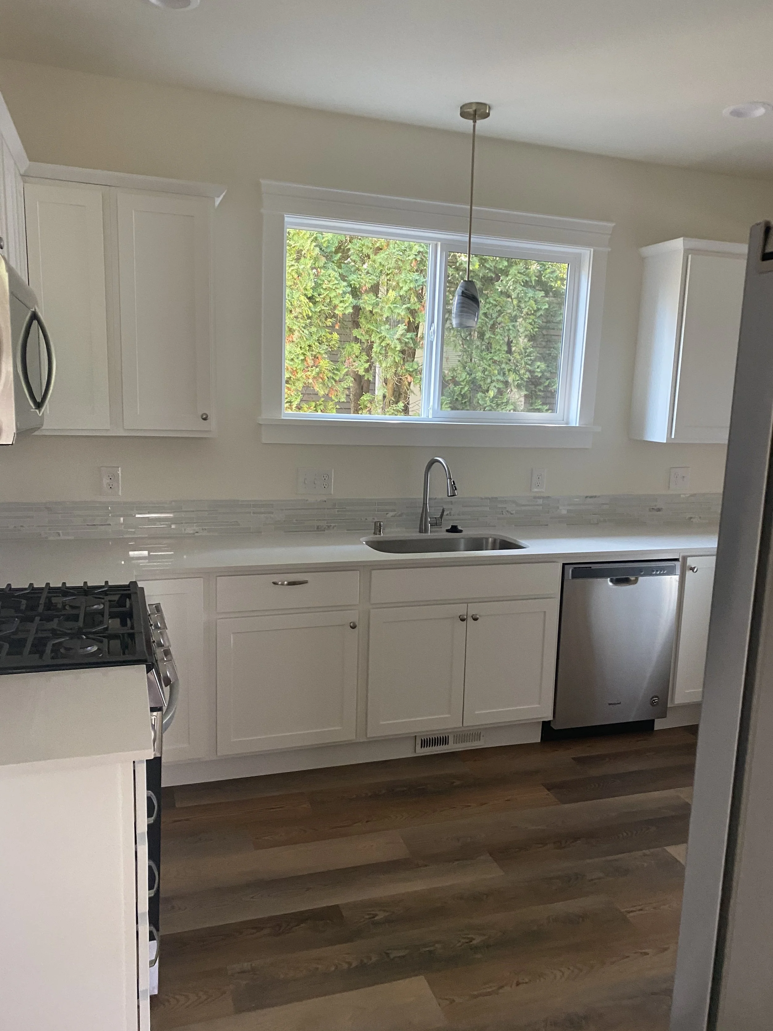 A clean modern kitchen with white cabinets and a window overlooking greenery. It features a stainless steel sink, a dishwasher, a stove, and a hanging pendant light.