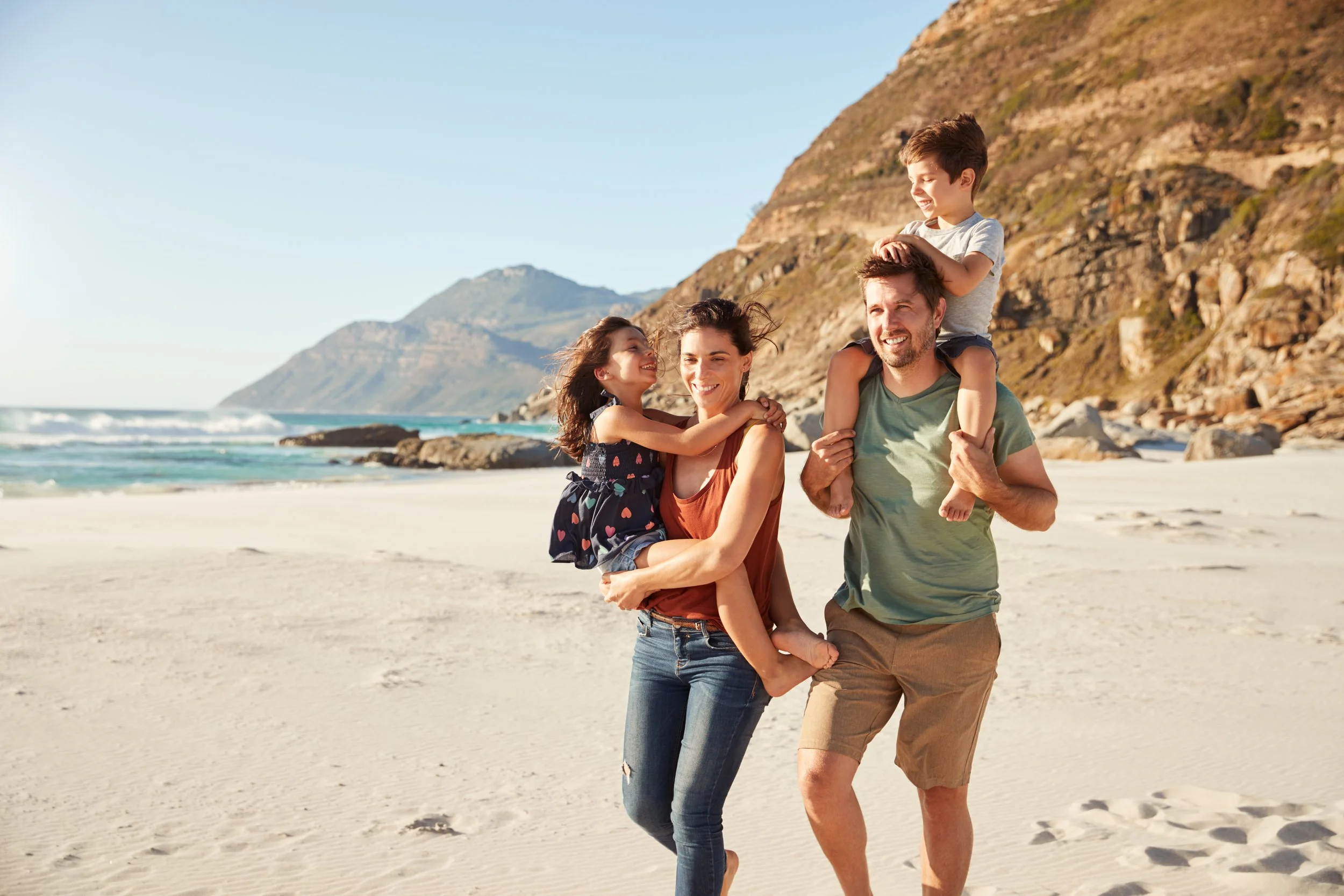 Family of four walking on the beach, smiling, with mountains in the background and the ocean waves nearby.