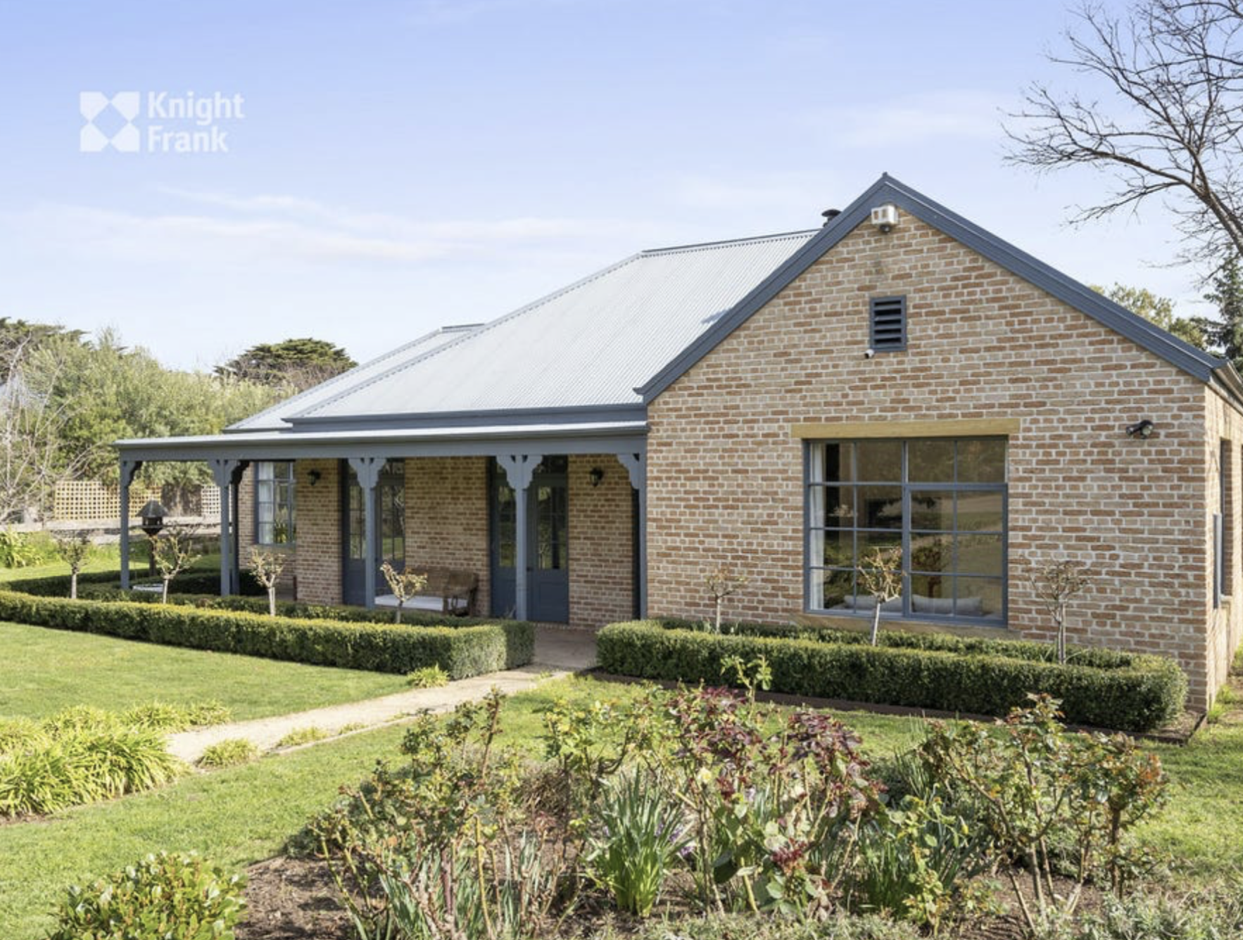 A brick house with a metal roof, large window, and front porch, surrounded by a well-maintained garden with shrubs, flowers, and trimmed hedges.