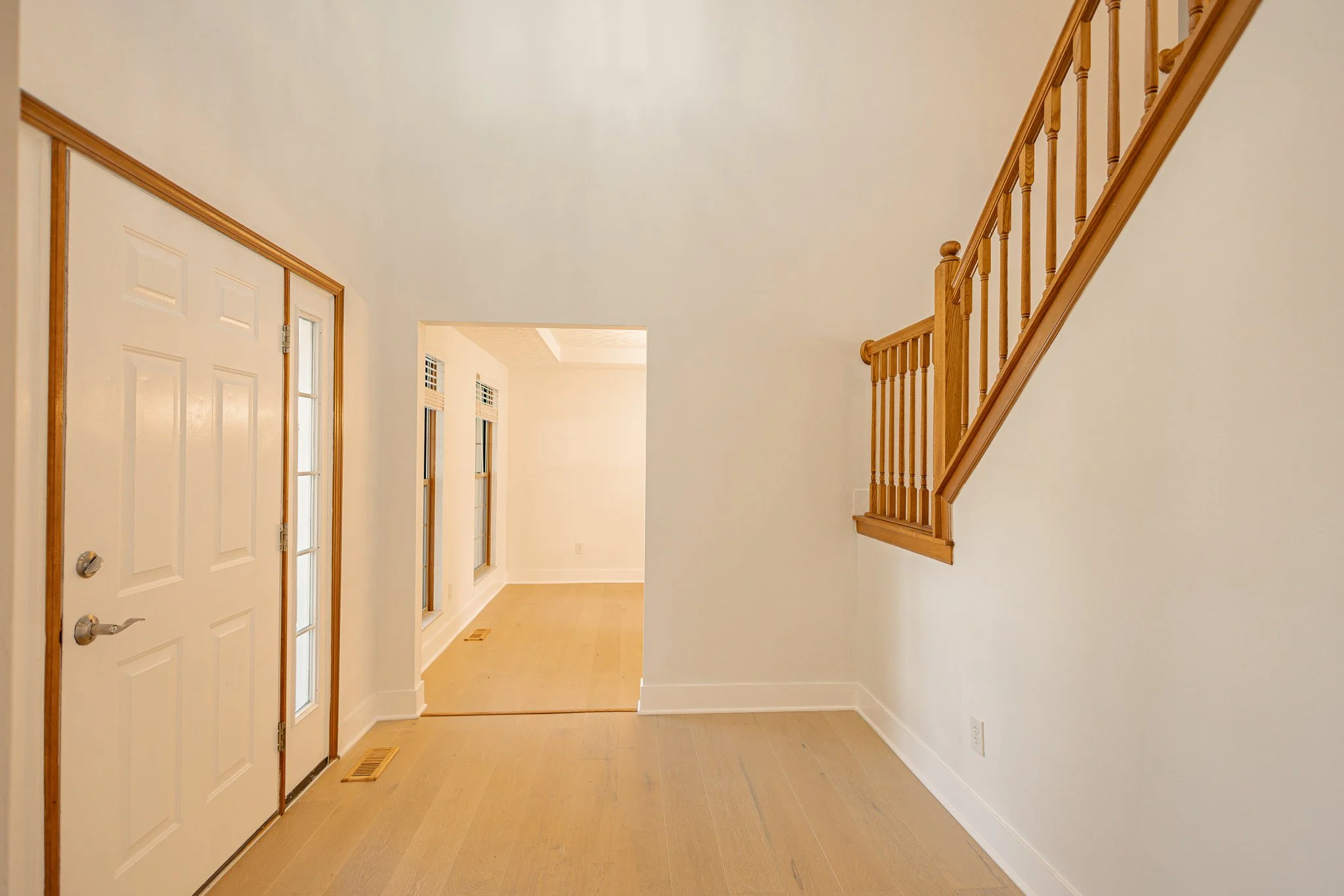 Interior view of a hallway with a front door on the left, featuring a small window, white walls, hardwood flooring, and a staircase with a wooden railing on the right.