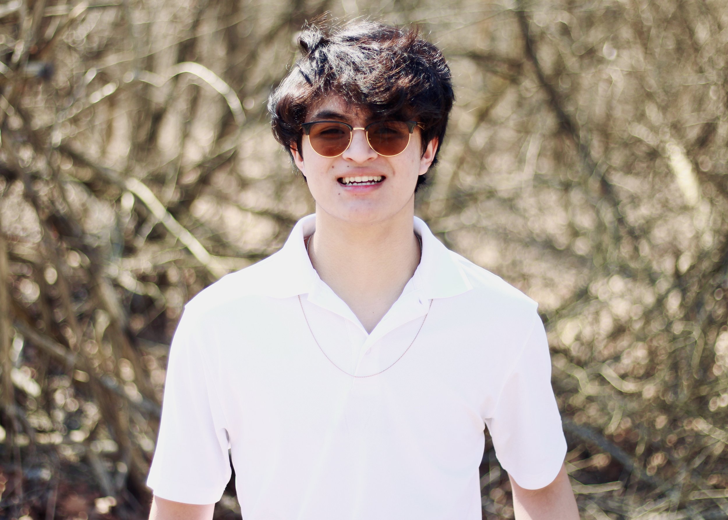 Person wearing sunglasses and a white shirt standing outdoors with trees in the background.