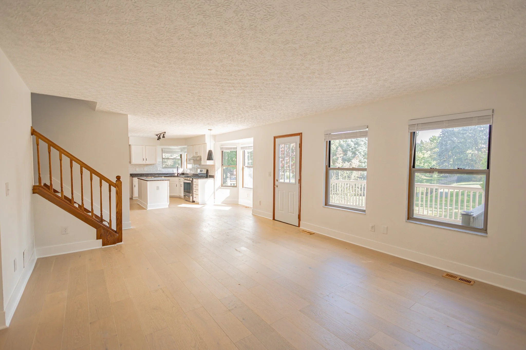 Empty living room and kitchen area with large windows, light wood flooring, white walls, and a staircase railing on the left.
