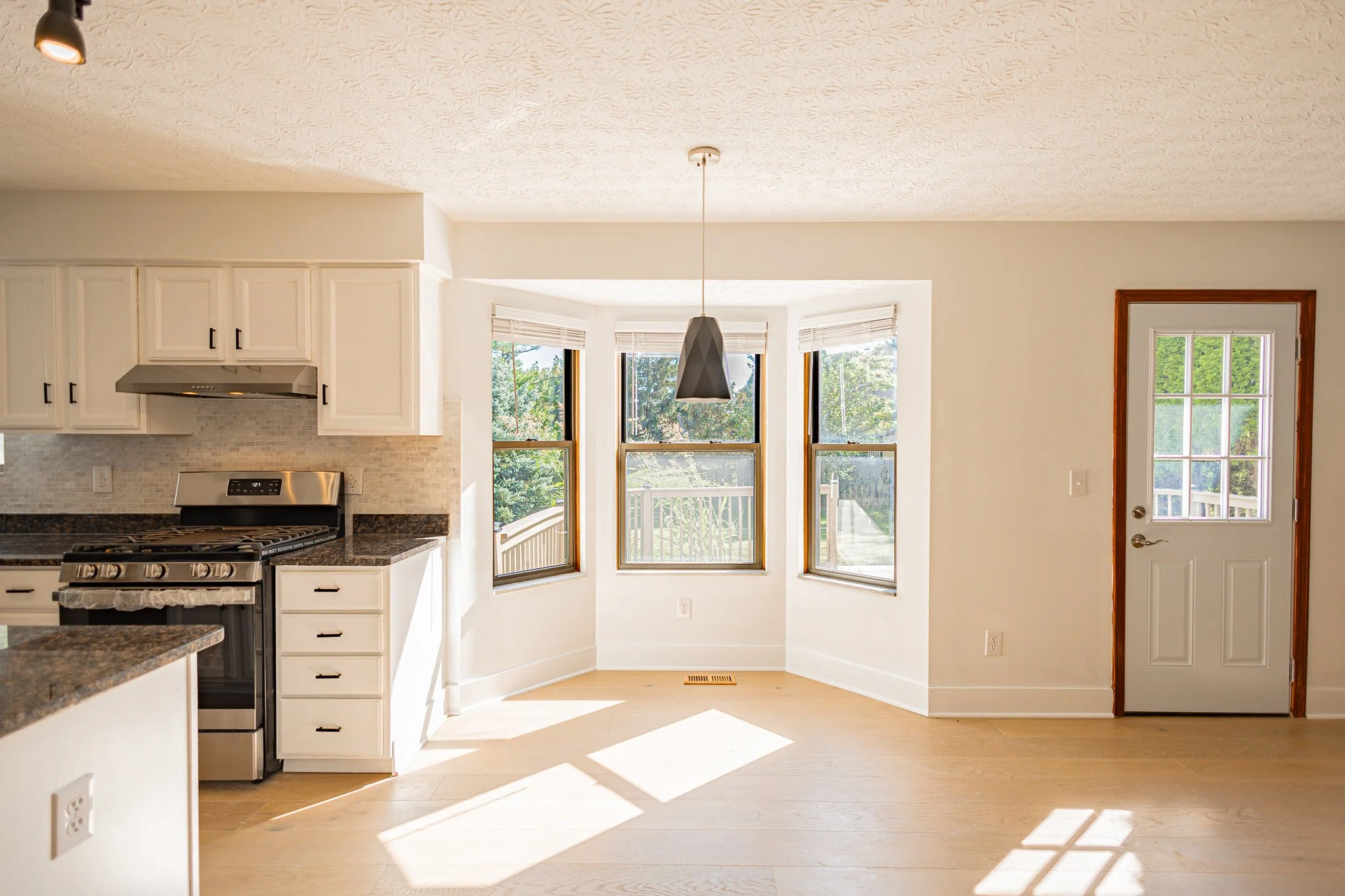Bright kitchen with white cabinets, black countertops, stainless steel stove, large bay window with blinds, and a door with a window, letting in natural light.