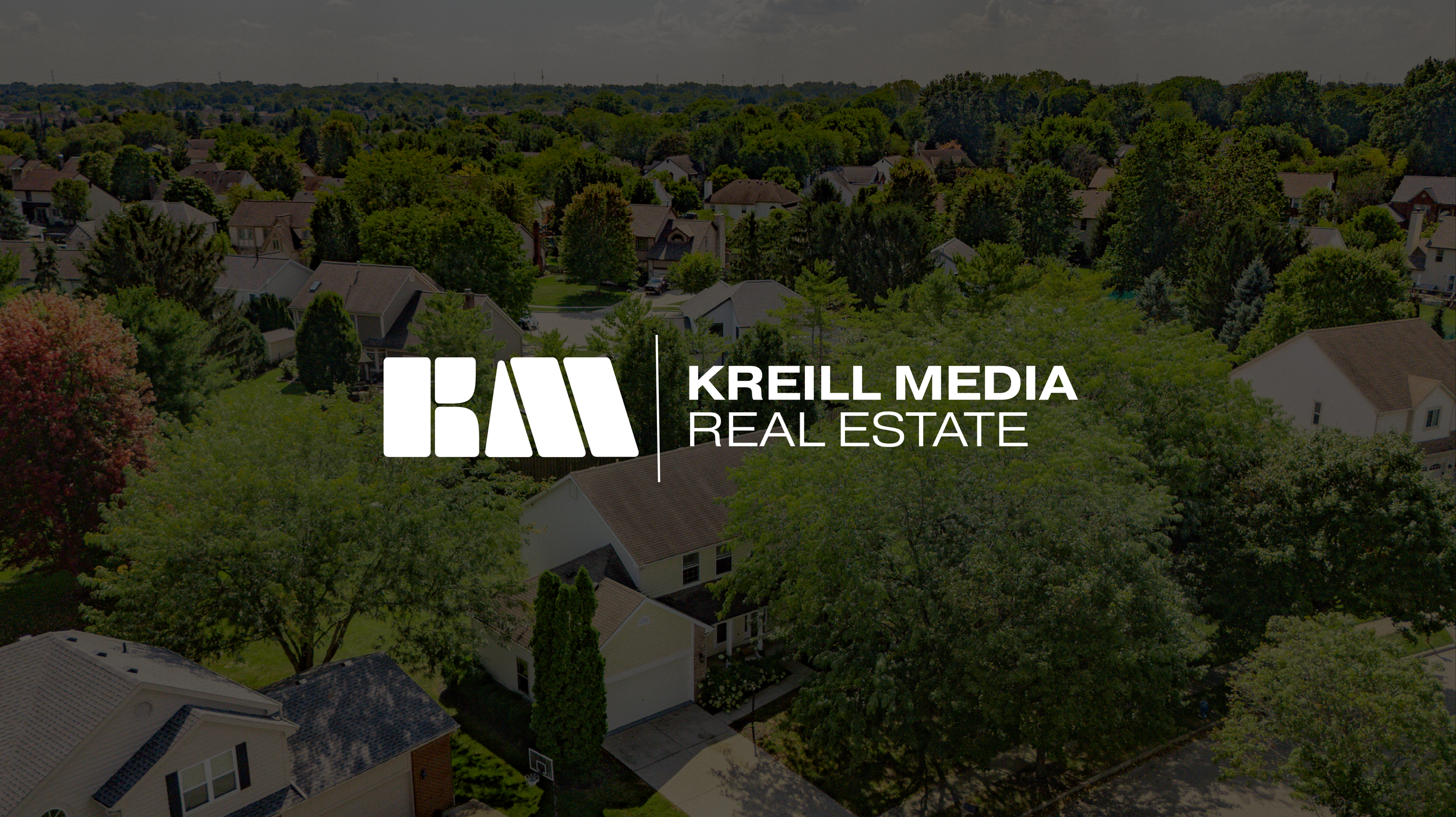 Aerial view of a suburban neighborhood with many houses, trees, and greenery under a cloudy sky. Overlayed text reads 'KREILL MEDIA REAL ESTATE' with a logo.