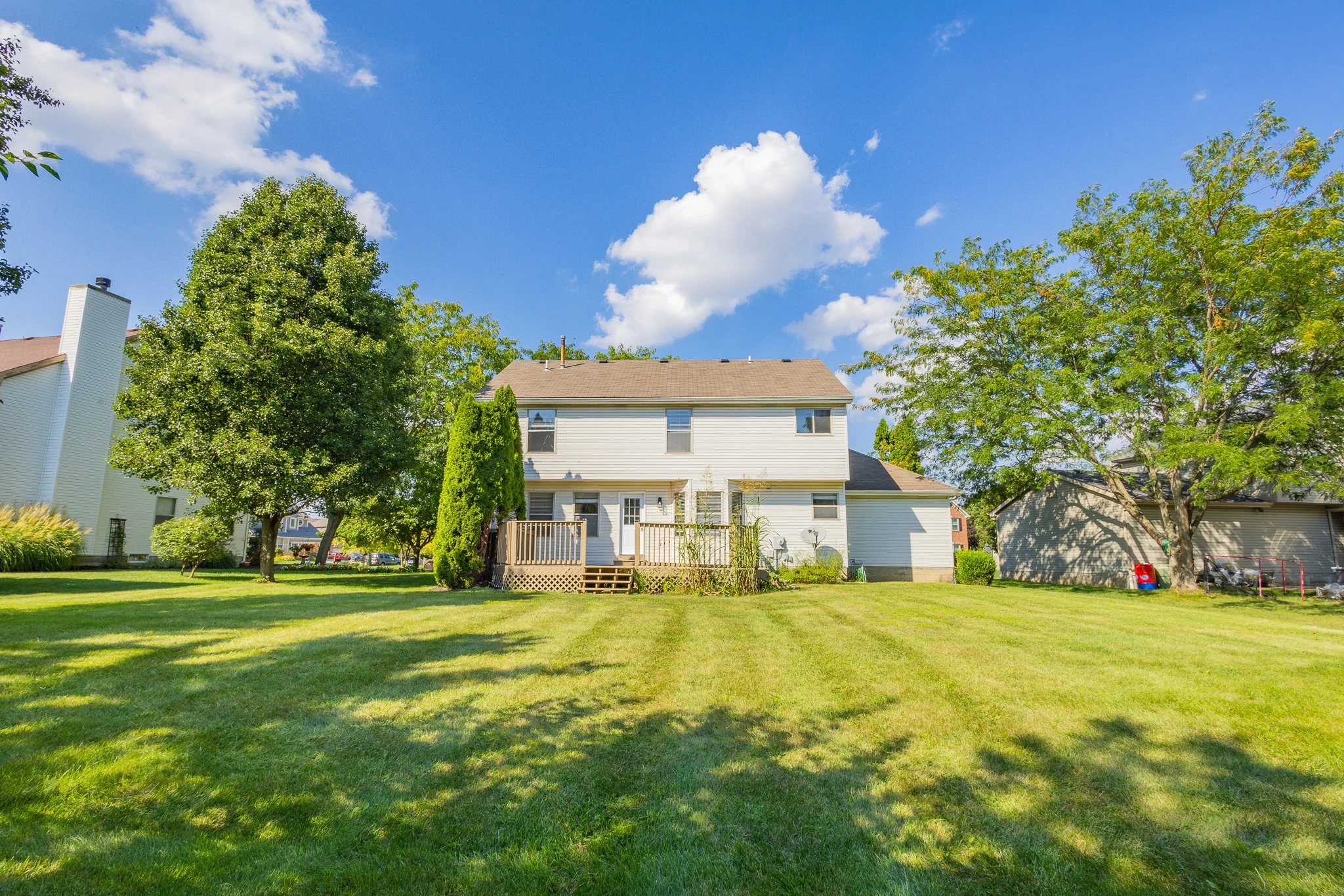 Backyard with a house, green lawn, trees, and a partly cloudy blue sky.