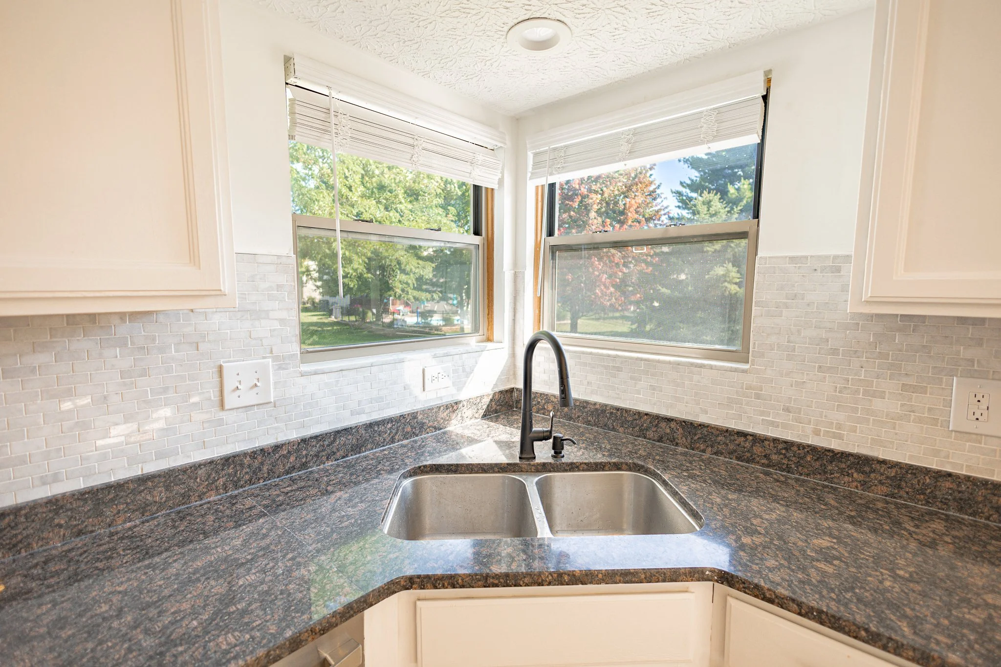 Kitchen with white cabinets, beige-tiled backsplash, black faucet, dark granite countertop, and large corner window with white blinds showing trees outside.
