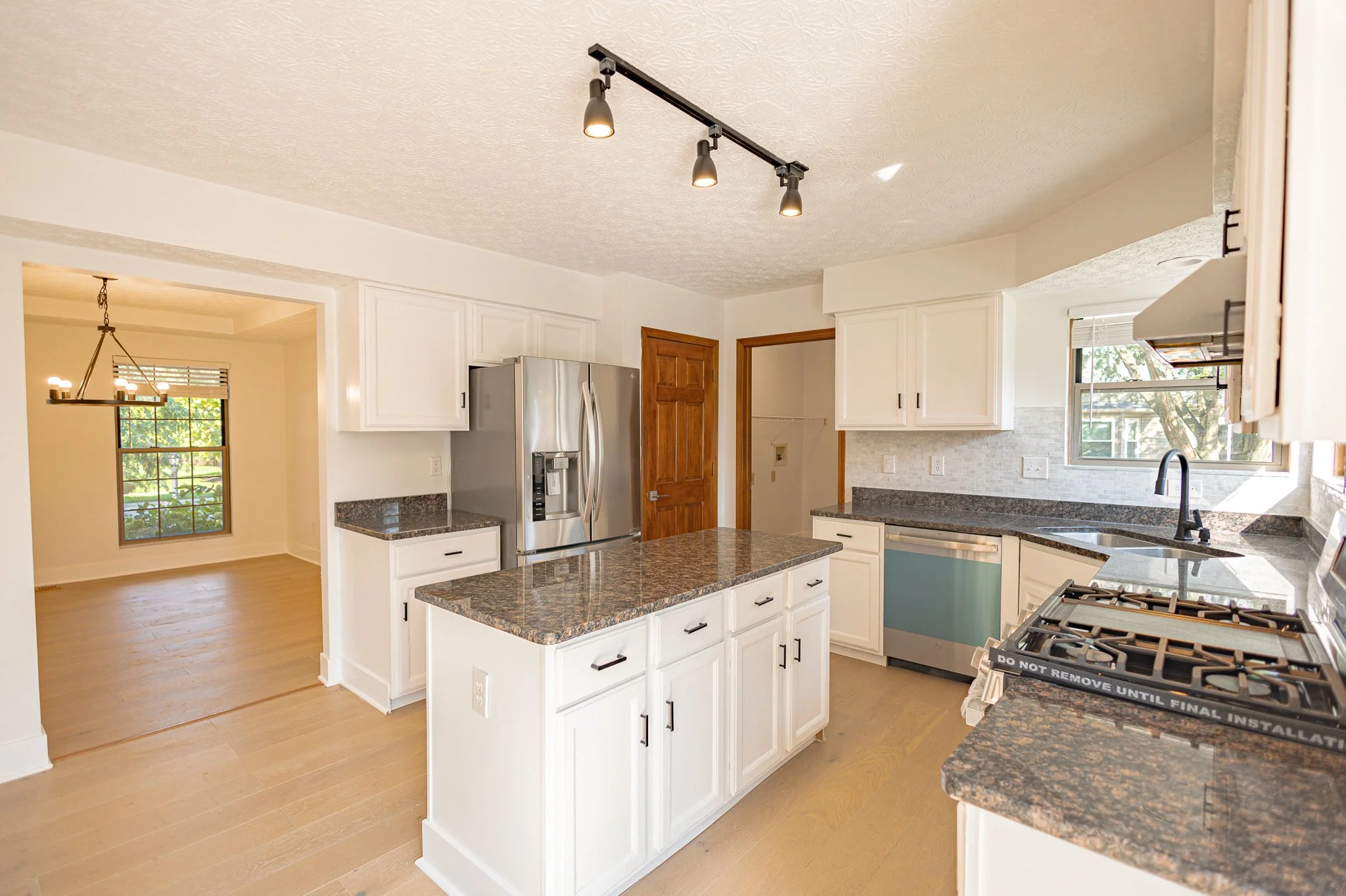 Kitchen with white cabinets, granite countertops, stainless steel refrigerator, and a gas stove. There is a small island and a window with a view of trees outside. A separate dining room with a chandelier is visible through a doorway.
