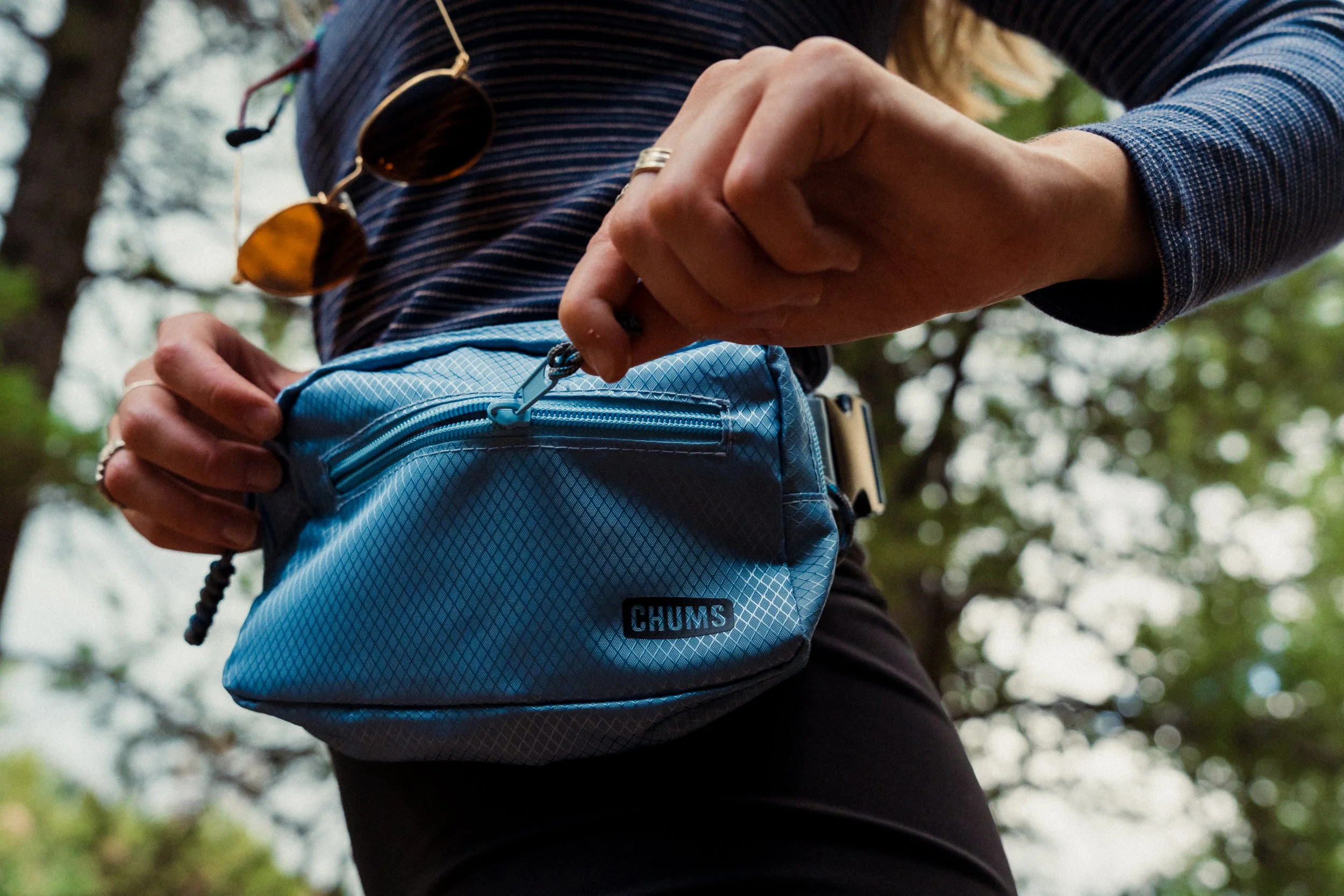 A person wearing a dark striped long sleeve shirt and black pants, holding a blue quilted Chums brand backpack with a zipper, with a brown leather strap visible, outdoors with trees and blurred green foliage in the background.