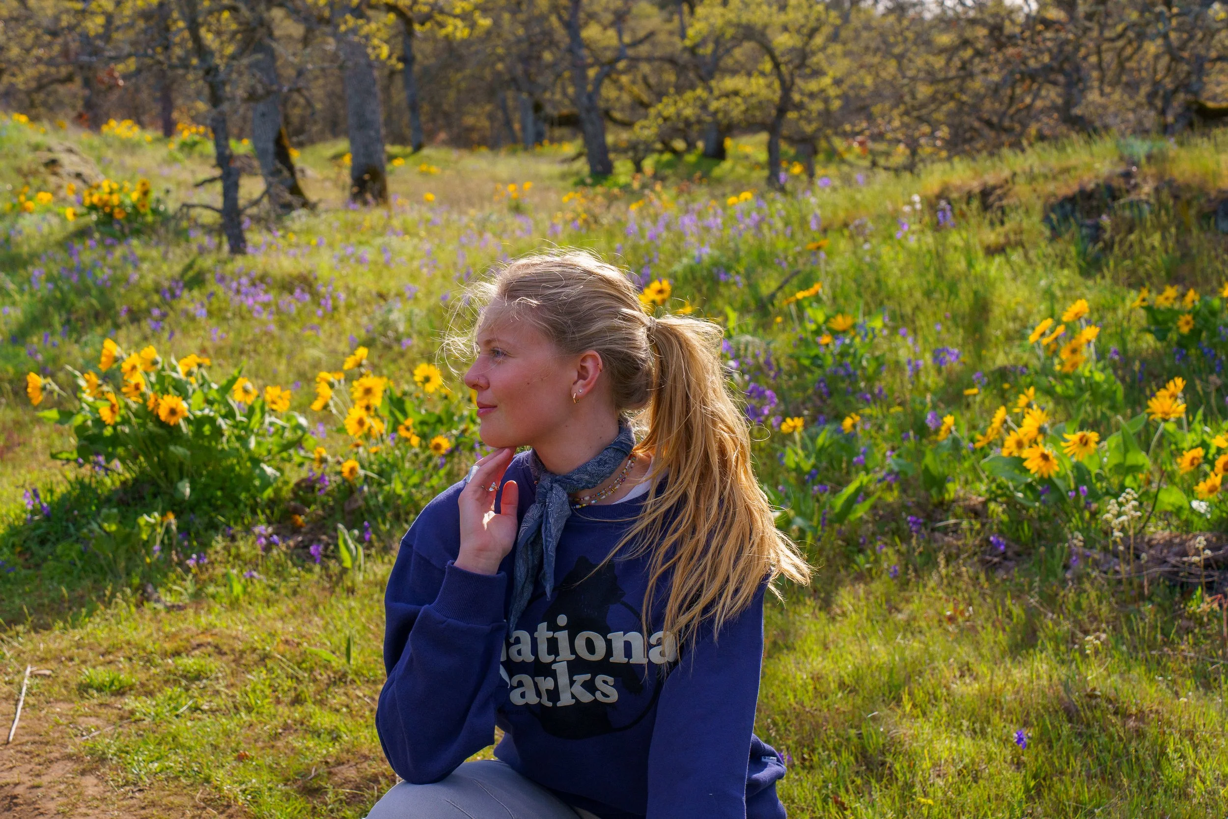 A young girl with long blonde hair in a ponytail, wearing a navy blue sweatshirt with white text, poses outdoors in a field of yellow and purple wildflowers with trees in the background on a sunny day.